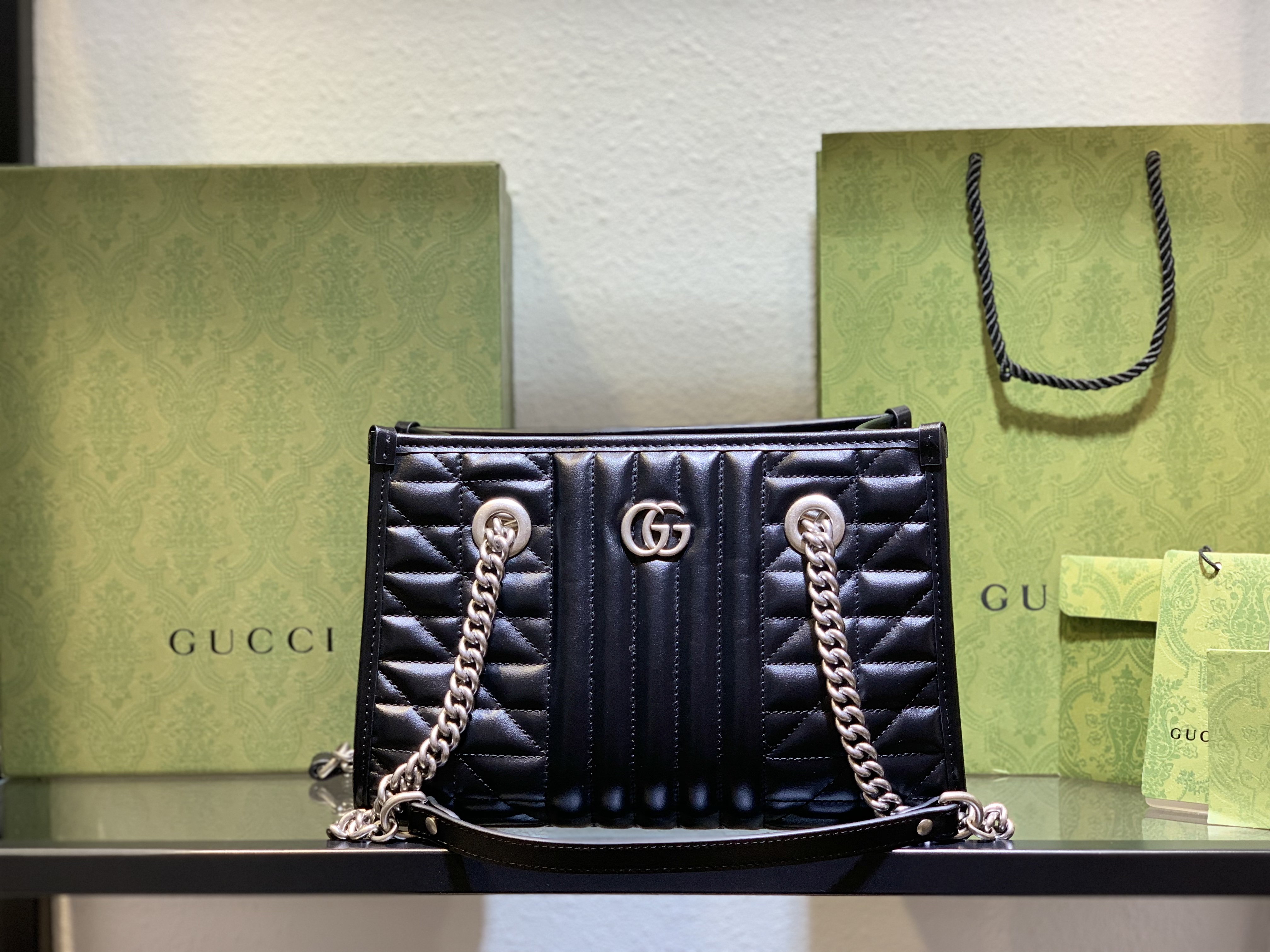 Gucci Black GG Marmont Quilted Shoulder Bag with silver GG logo and chain strap, displayed with Gucci packaging.