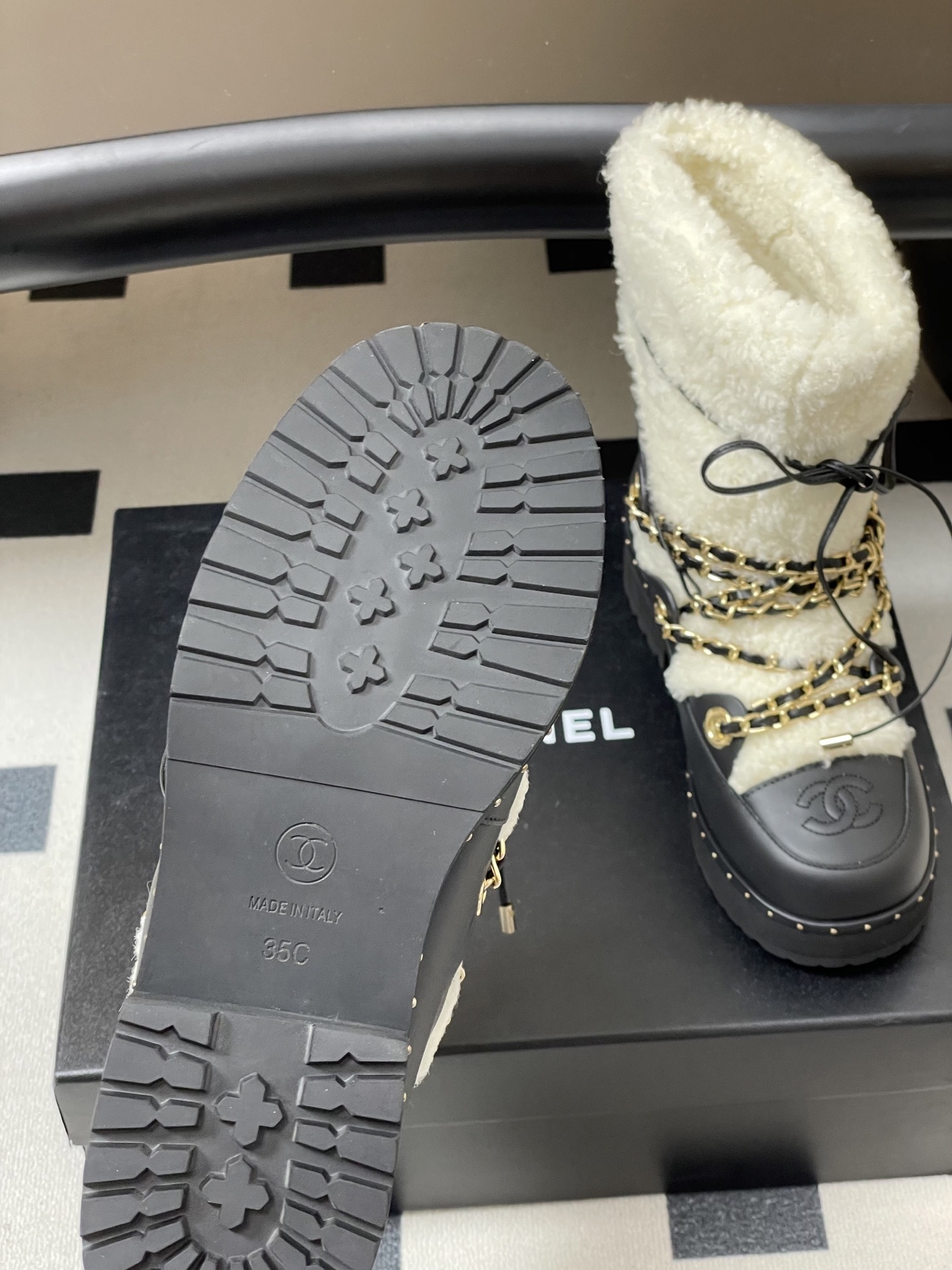 Pair of Chanel black leather and white shearling winter boots with gold chain detailing and embossed CC logo on a black table. - Hình 8