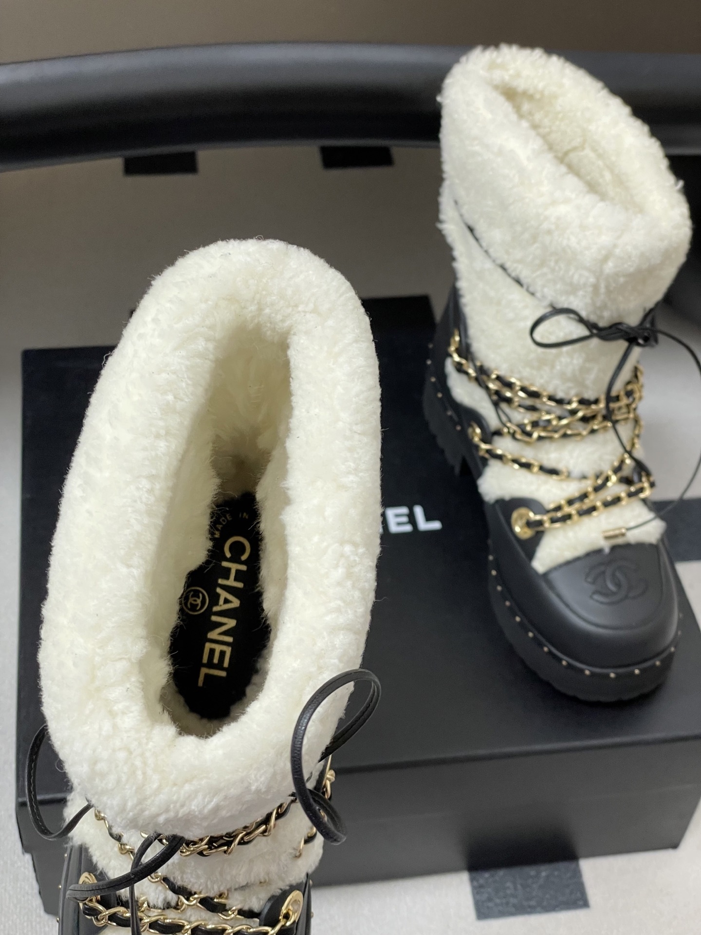 Pair of Chanel black leather and white shearling winter boots with gold chain detailing and embossed CC logo on a black table. - Hình 6