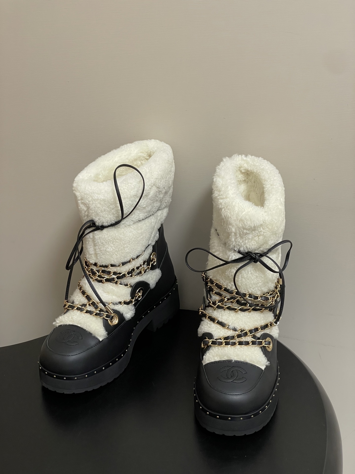 Pair of Chanel black leather and white shearling winter boots with gold chain detailing and embossed CC logo on a black table. - Hình 3