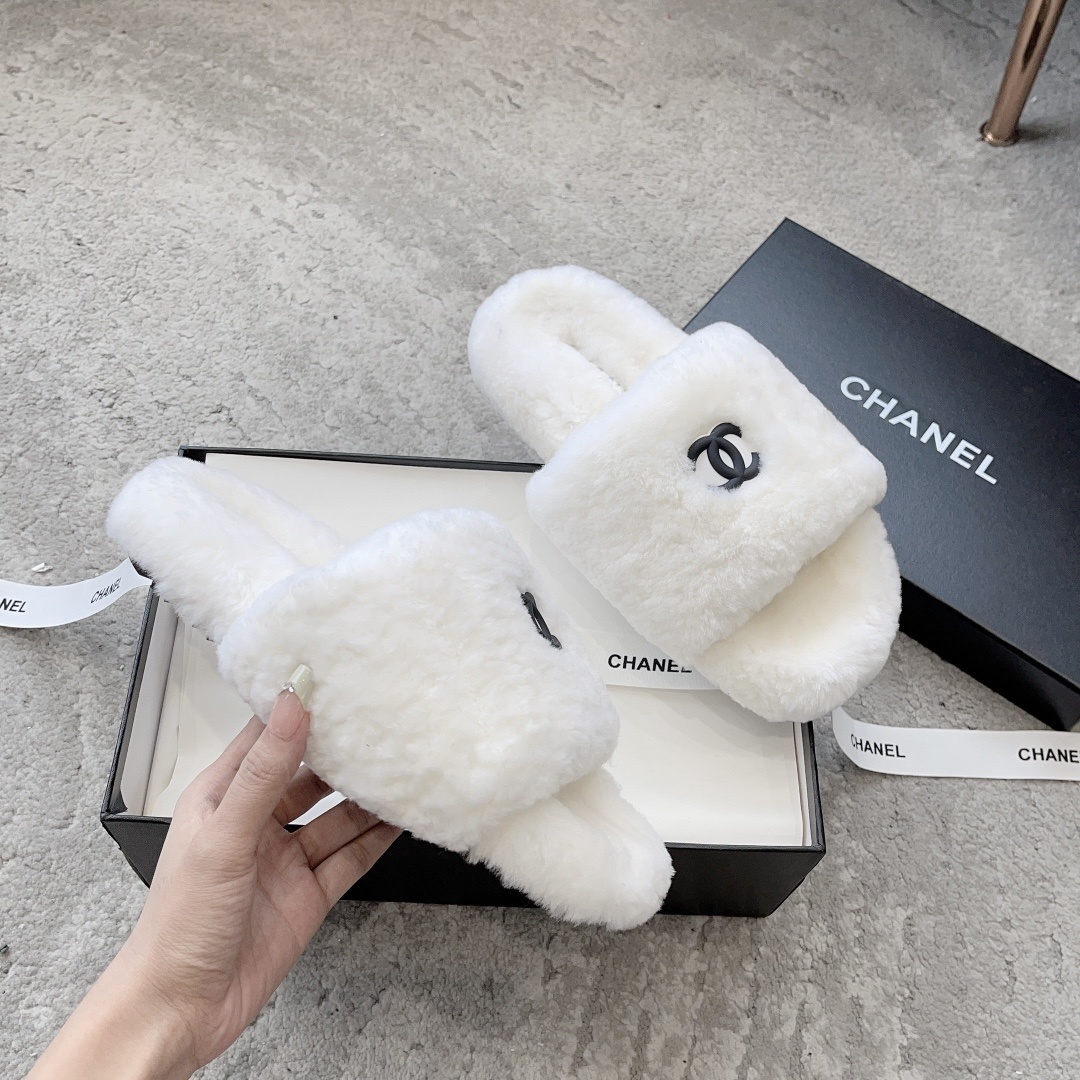 A pair of fluffy white faux fur slippers featuring a black interlaced logo, presented on a black designer box. - Hình 3