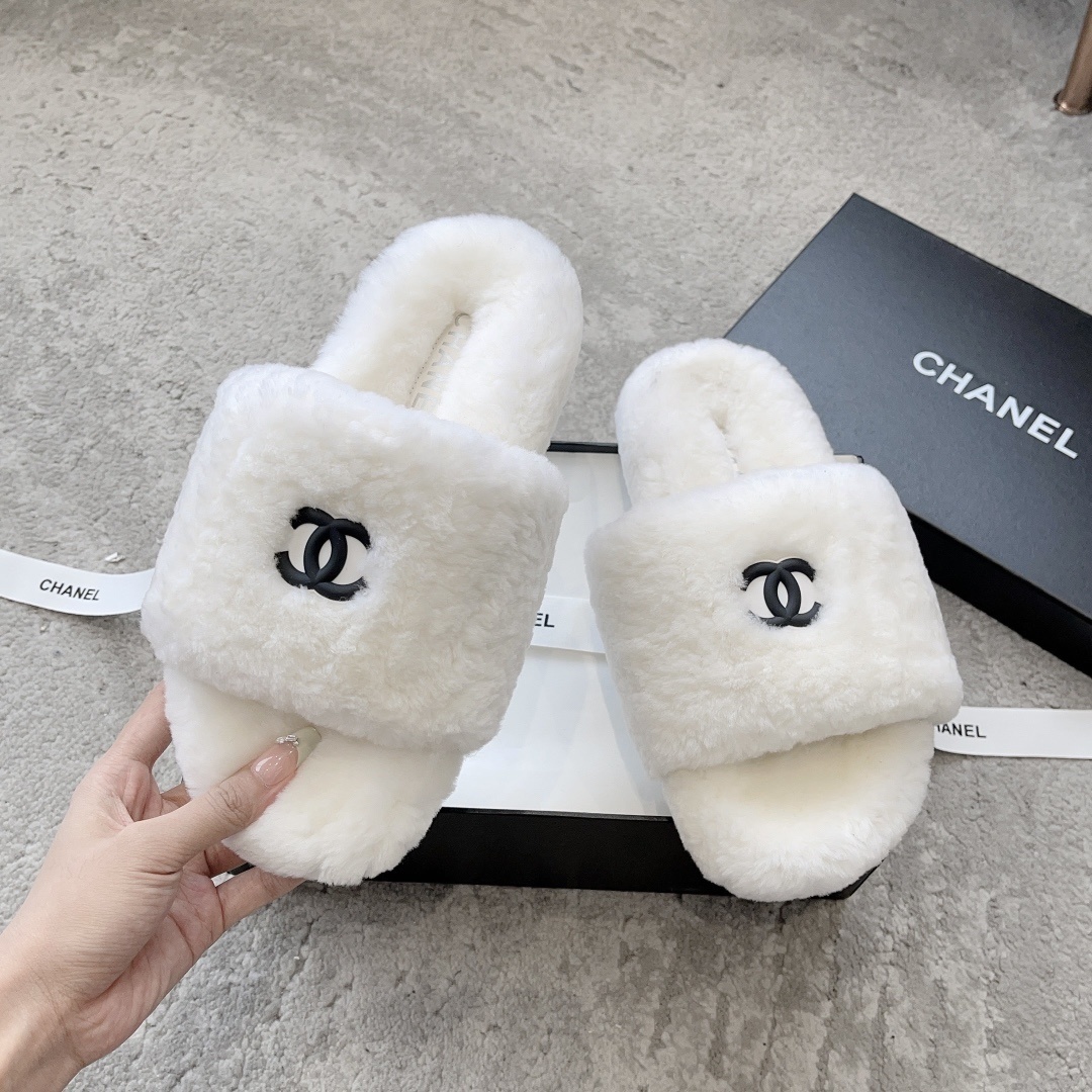 A pair of fluffy white faux fur slippers featuring a black interlaced logo, presented on a black designer box. - Hình 2