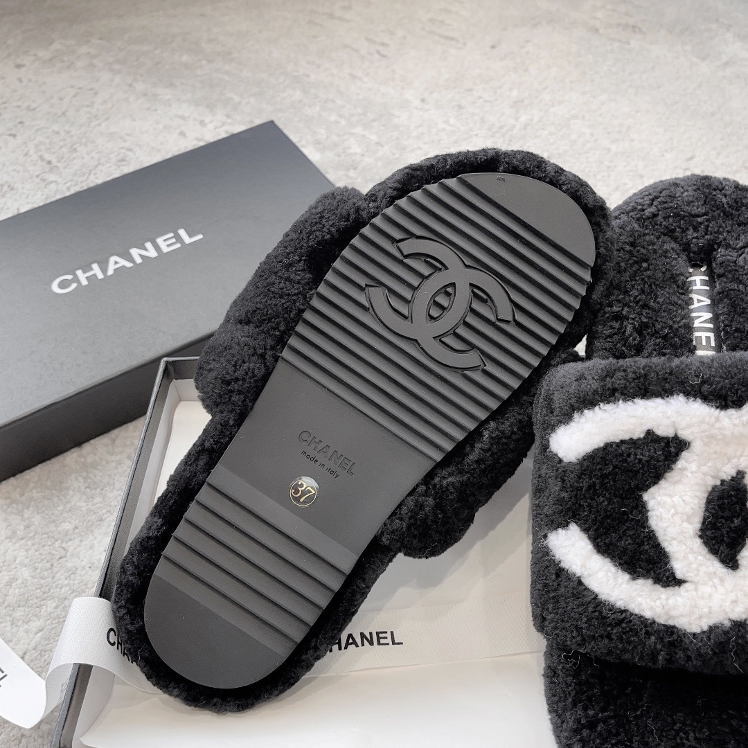 A pair of black fuzzy Chanel shearling slippers with a large white interlocking CC logo, presented in a black Chanel box with a white ribbon. - Hình 9