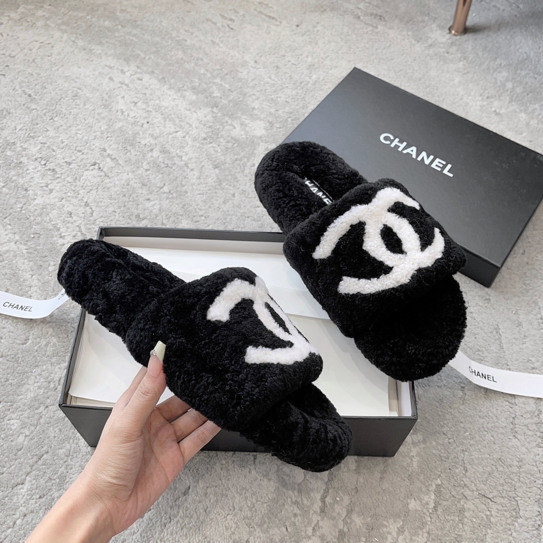 A pair of black fuzzy Chanel shearling slippers with a large white interlocking CC logo, presented in a black Chanel box with a white ribbon. - Hình 3