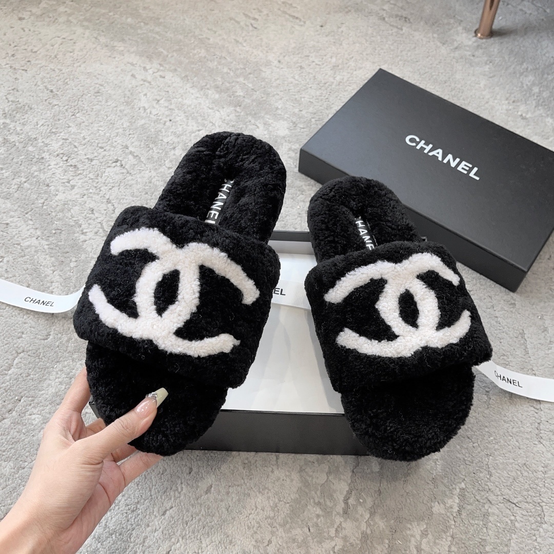 A pair of black fuzzy Chanel shearling slippers with a large white interlocking CC logo, presented in a black Chanel box with a white ribbon. - Hình 2