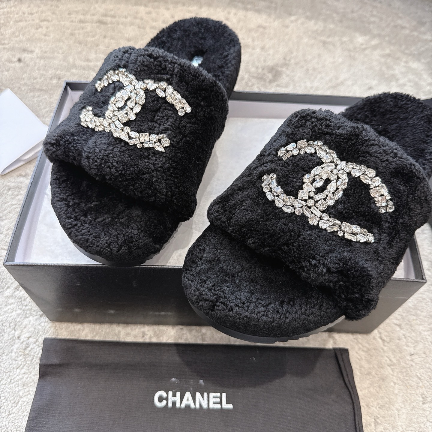 A pair of black fuzzy Chanel slides with large crystal interlocking CC logos on top, presented in a black brand box. - Hình 7
