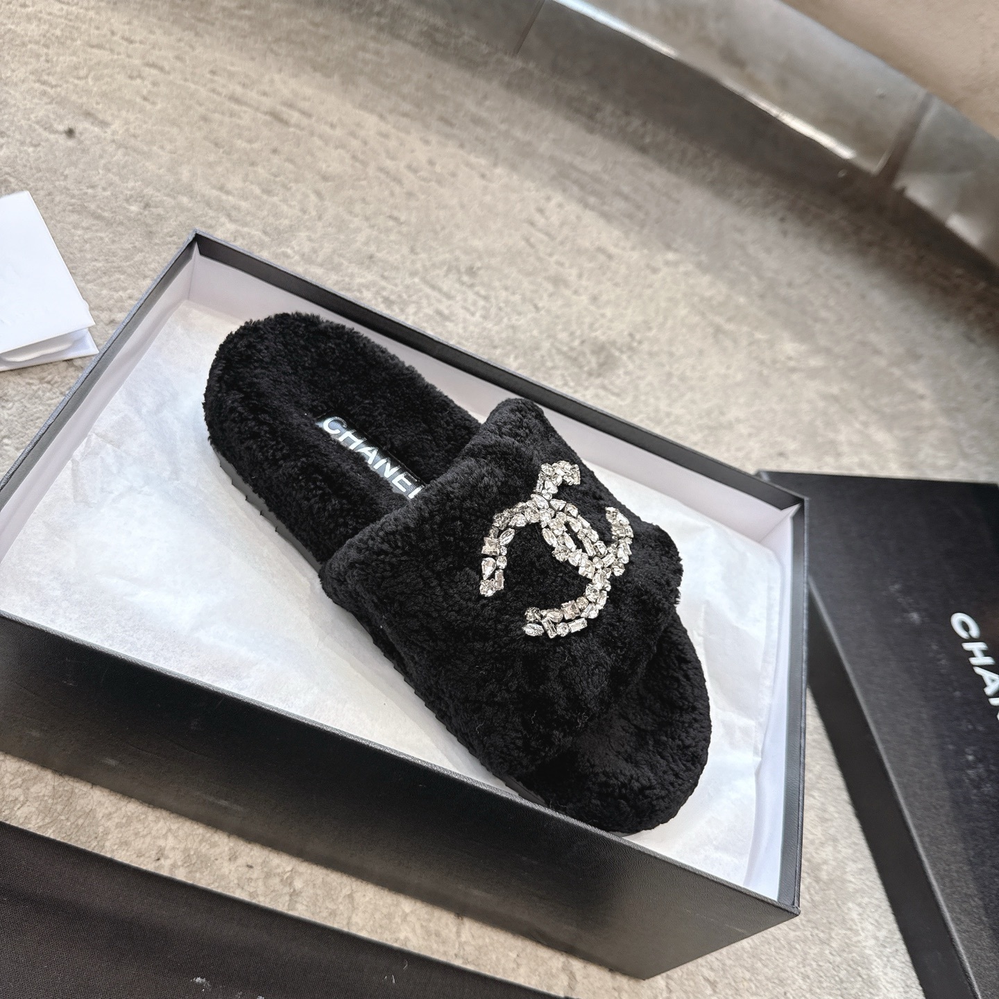 A pair of black fuzzy Chanel slides with large crystal interlocking CC logos on top, presented in a black brand box. - Hình 4