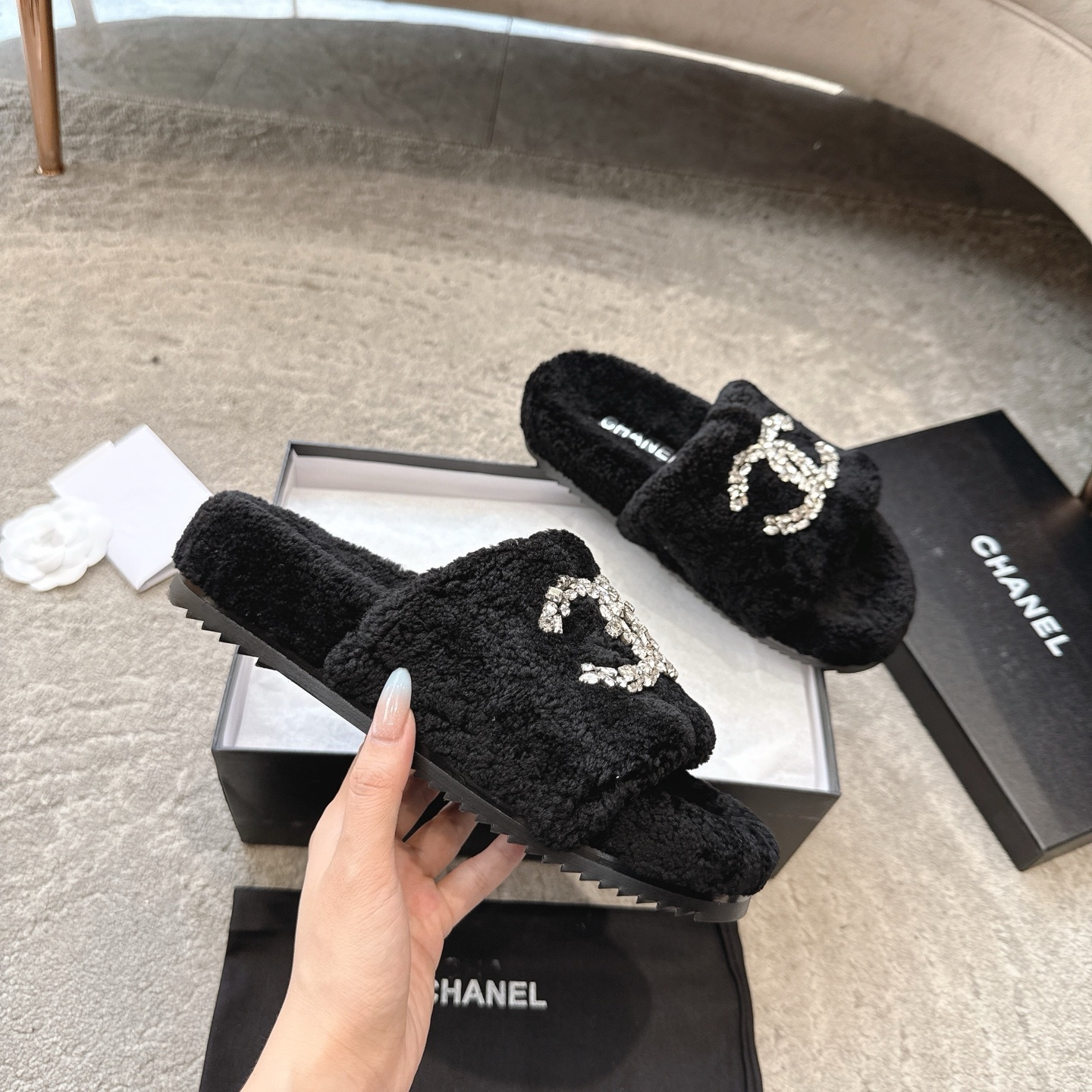 A pair of black fuzzy Chanel slides with large crystal interlocking CC logos on top, presented in a black brand box. - Hình 3