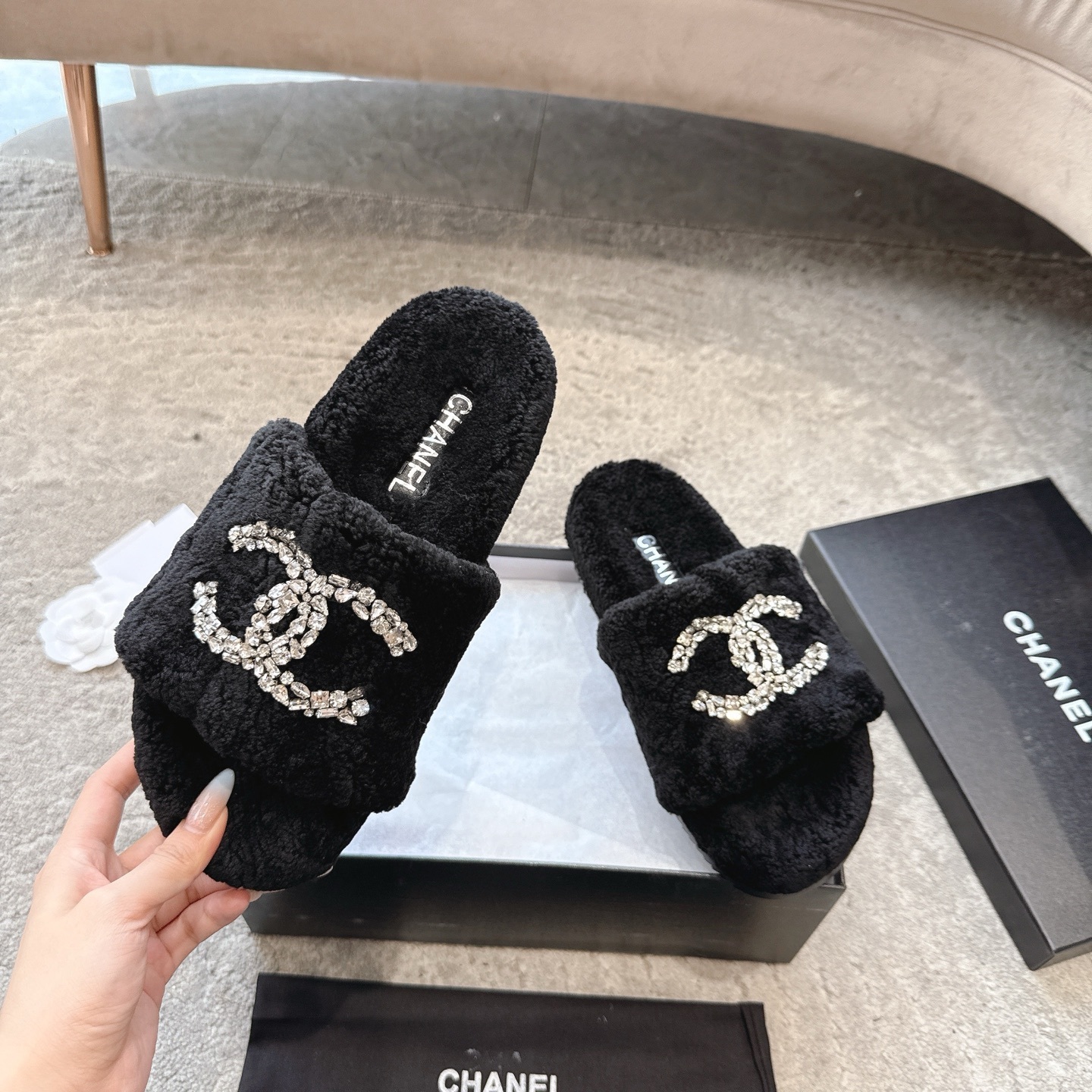 A pair of black fuzzy Chanel slides with large crystal interlocking CC logos on top, presented in a black brand box. - Hình 2