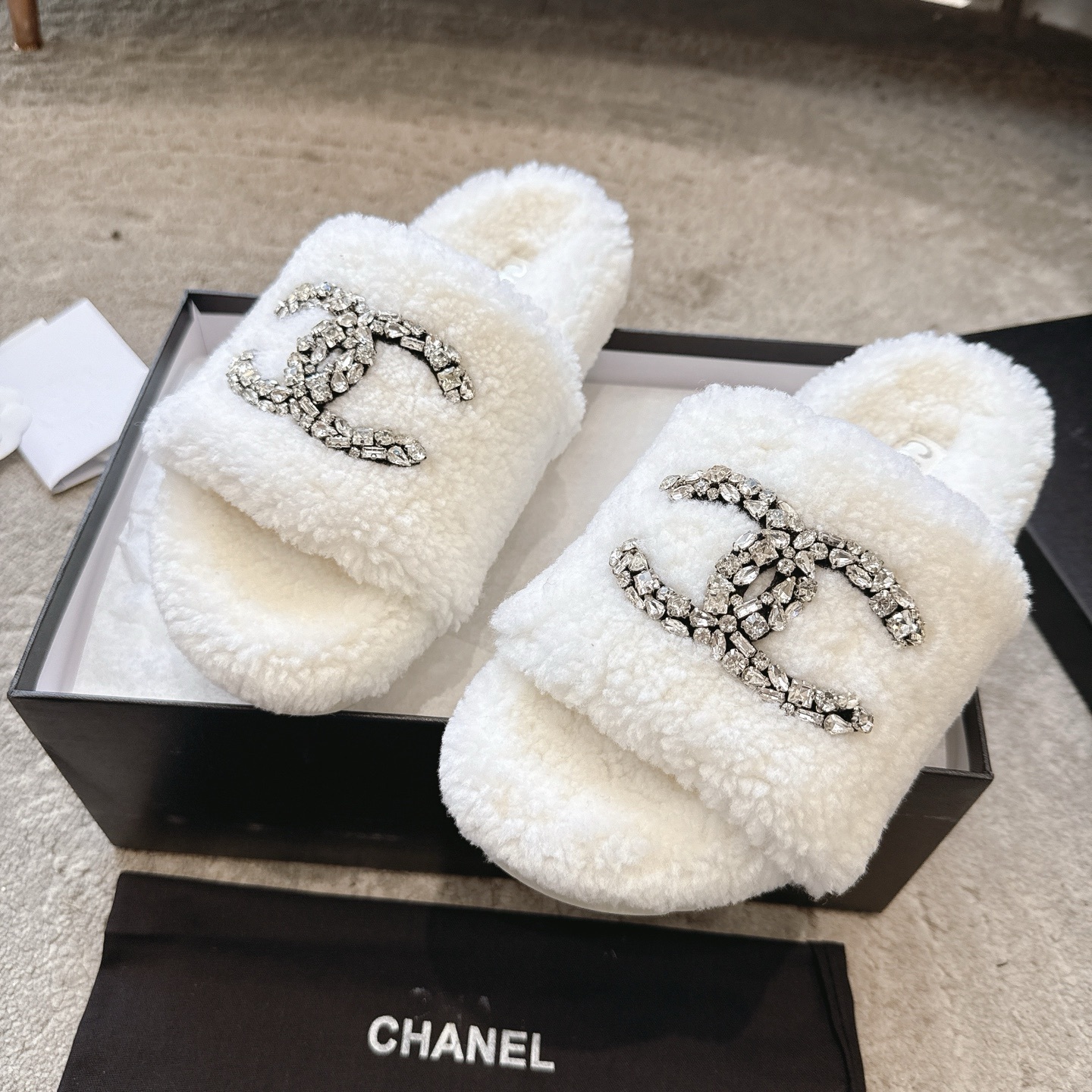 A pair of luxury white Chanel shearling slides with crystal-encrusted CC logos presented in a black brand box. - Hình 7