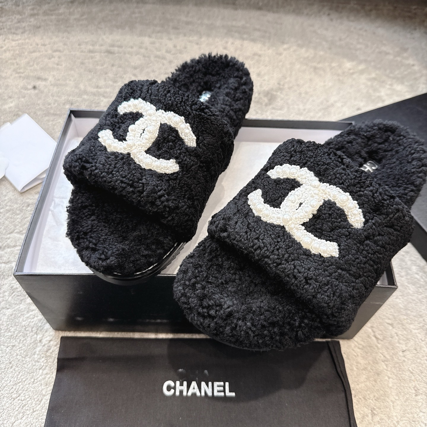 A pair of black fuzzy Chanel shearling slides with a large white pearl-embellished interlocking CC logo on the strap, shown in their original box. - Hình 7