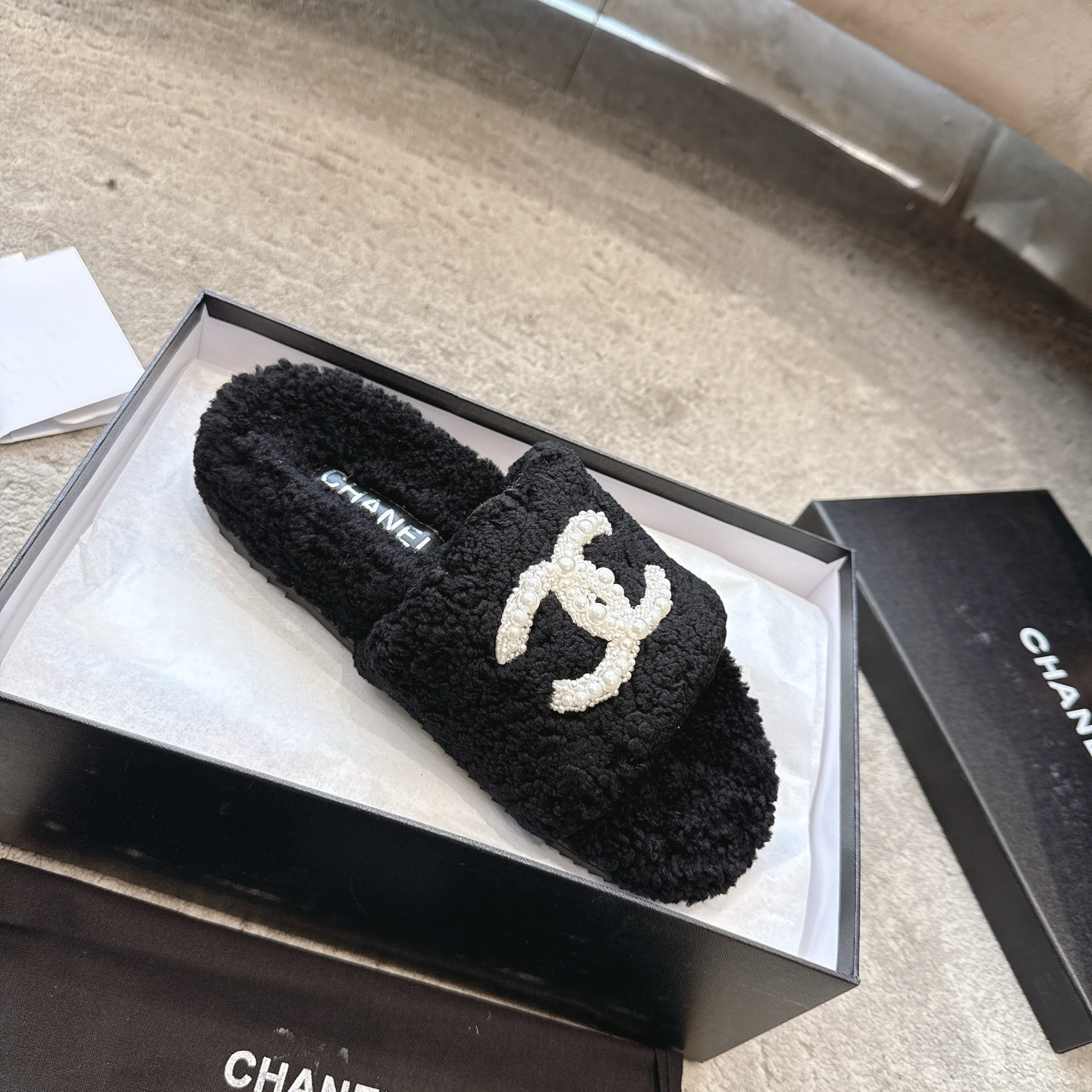 A pair of black fuzzy Chanel shearling slides with a large white pearl-embellished interlocking CC logo on the strap, shown in their original box. - Hình 4