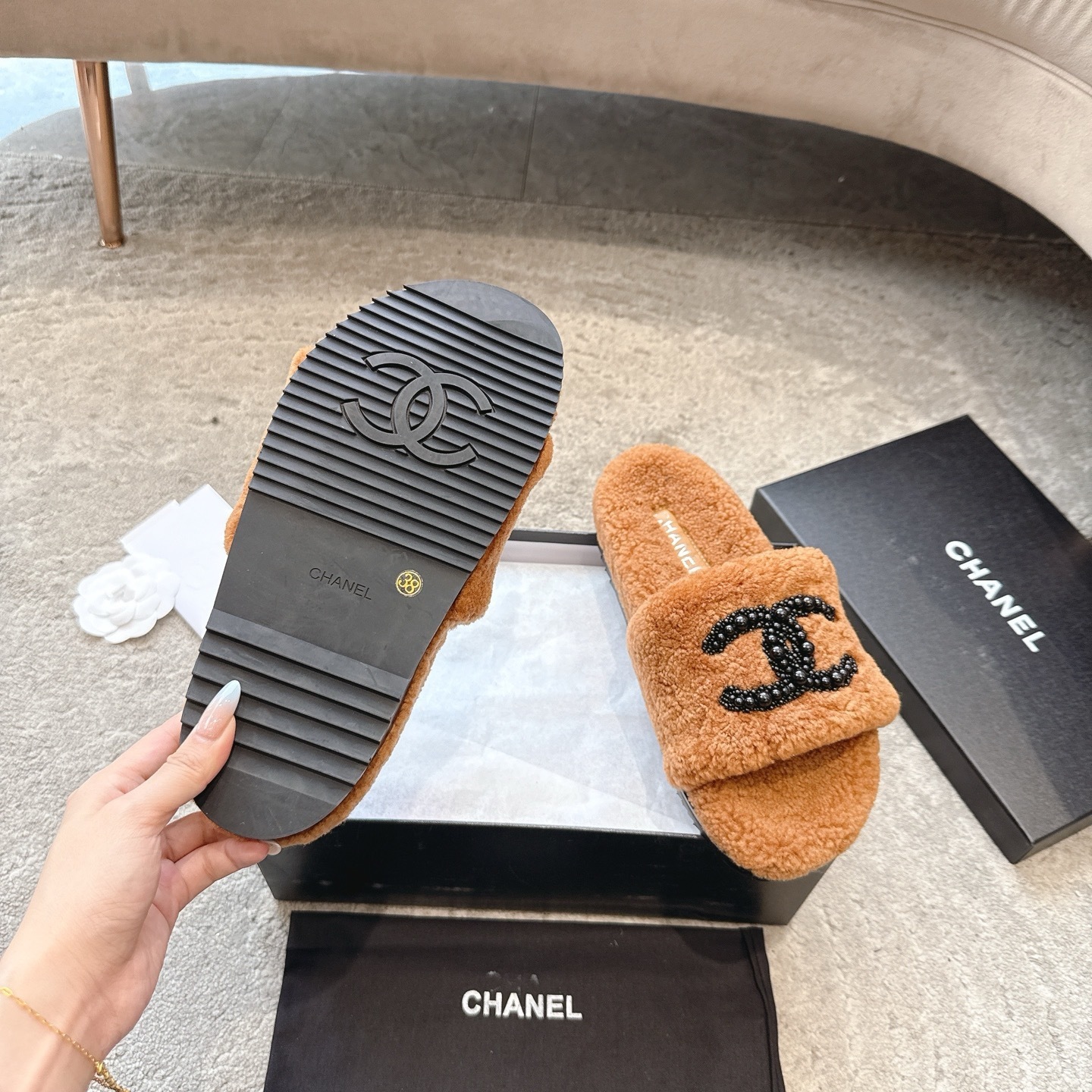 A pair of luxury brown shearling slides with black beaded Chanel CC logo sitting in a black designer box. - Hình 9