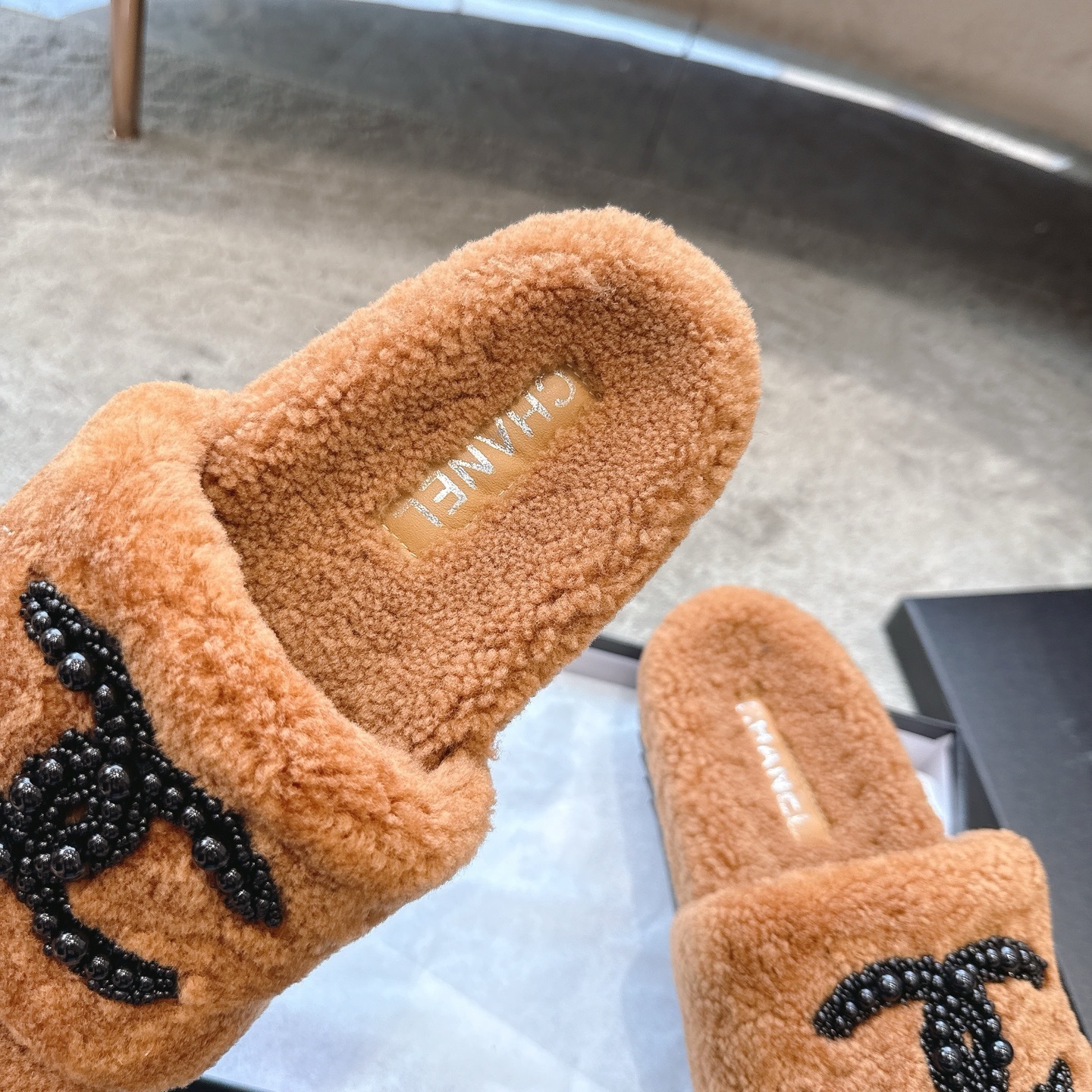 A pair of luxury brown shearling slides with black beaded Chanel CC logo sitting in a black designer box. - Hình 8