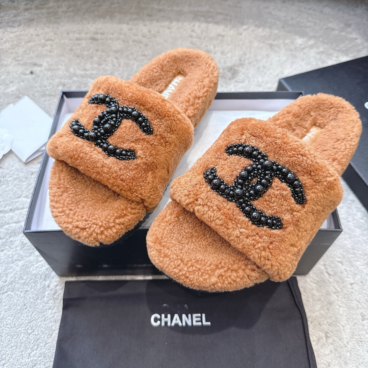A pair of luxury brown shearling slides with black beaded Chanel CC logo sitting in a black designer box. - Hình 7