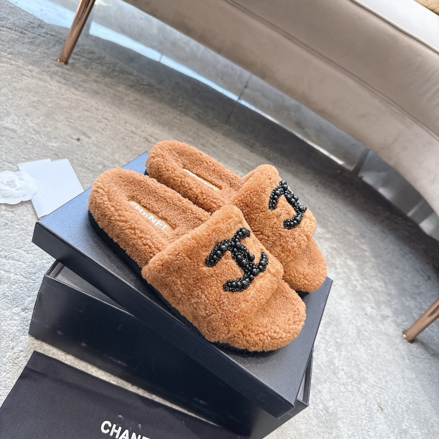 A pair of luxury brown shearling slides with black beaded Chanel CC logo sitting in a black designer box. - Hình 6