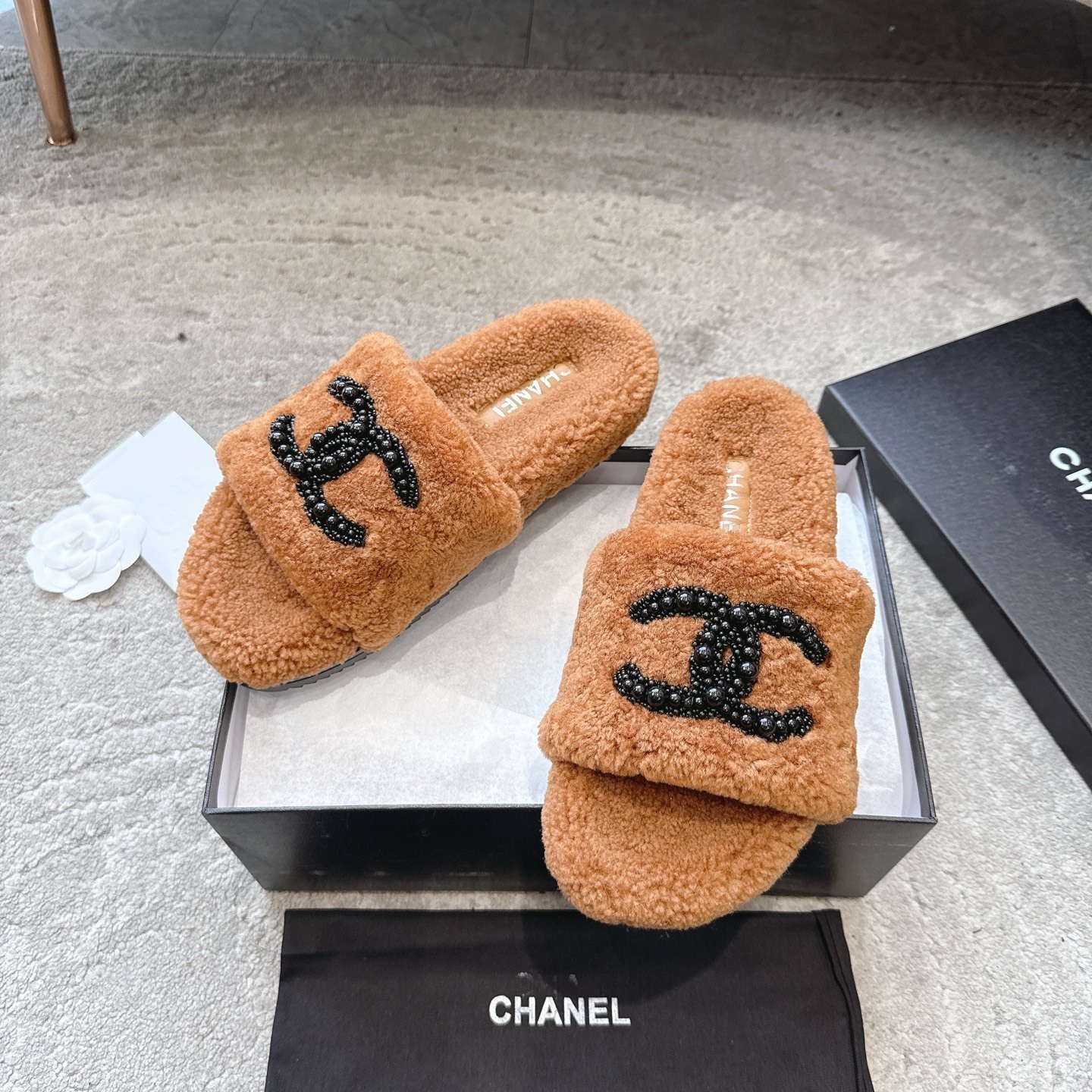 A pair of luxury brown shearling slides with black beaded Chanel CC logo sitting in a black designer box. - Hình 5