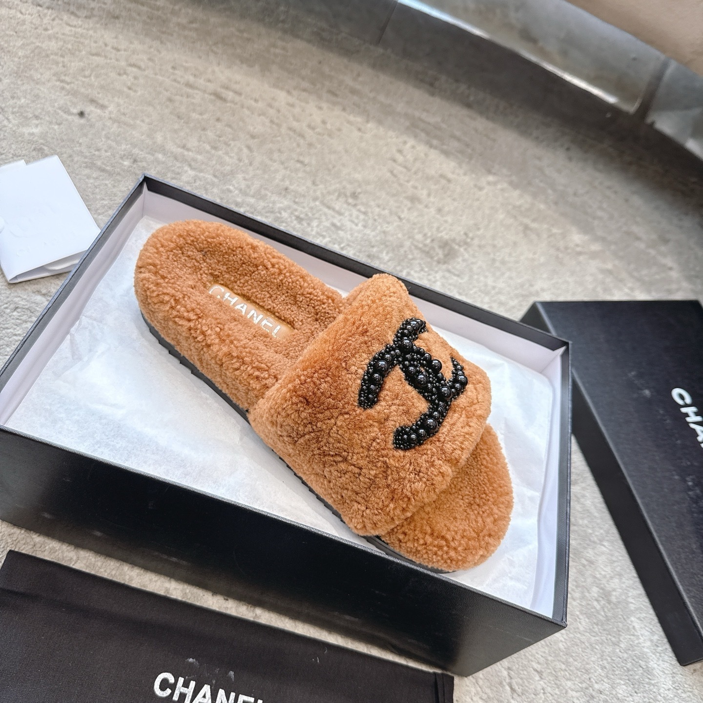 A pair of luxury brown shearling slides with black beaded Chanel CC logo sitting in a black designer box. - Hình 4