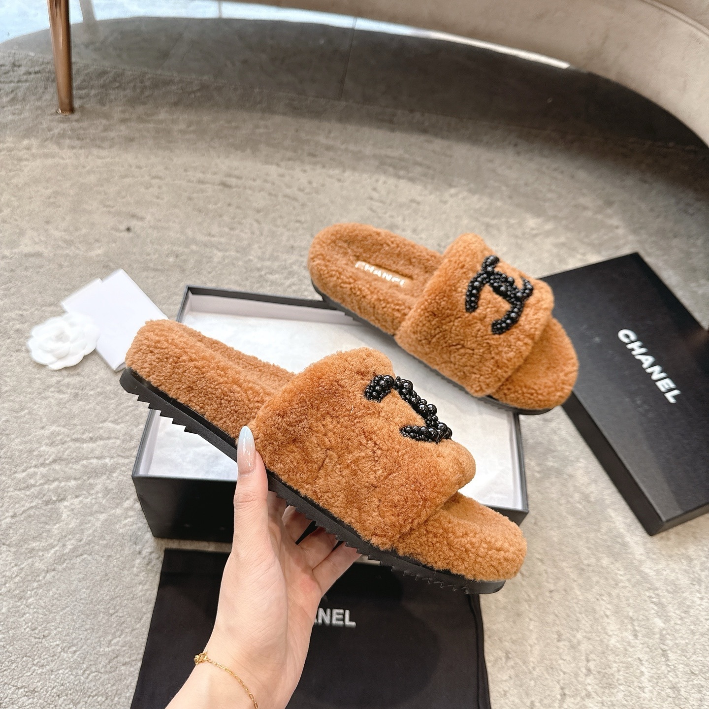 A pair of luxury brown shearling slides with black beaded Chanel CC logo sitting in a black designer box. - Hình 3