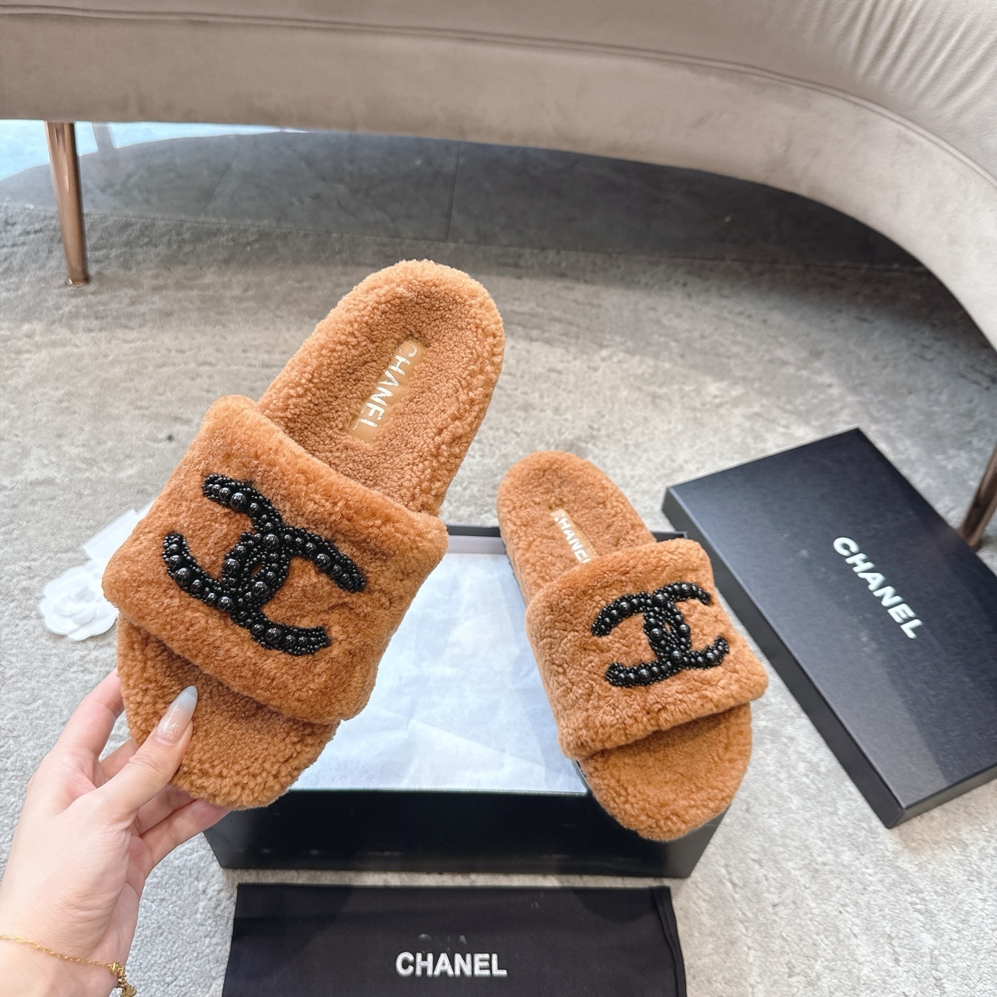 A pair of luxury brown shearling slides with black beaded Chanel CC logo sitting in a black designer box. - Hình 2