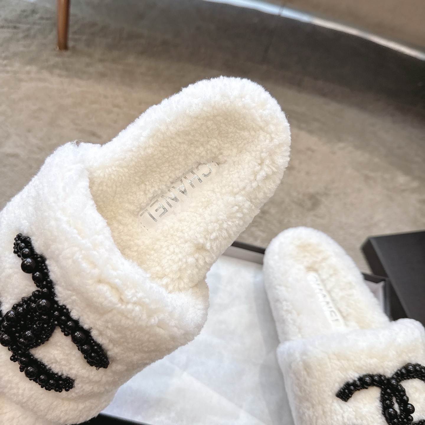 A pair of luxury Chanel white shearling slides featuring a black beaded CC logo, presented in a black Chanel box with a dust bag. - Hình 8