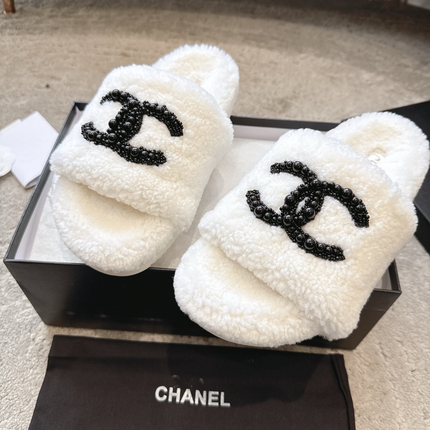 A pair of luxury Chanel white shearling slides featuring a black beaded CC logo, presented in a black Chanel box with a dust bag. - Hình 7