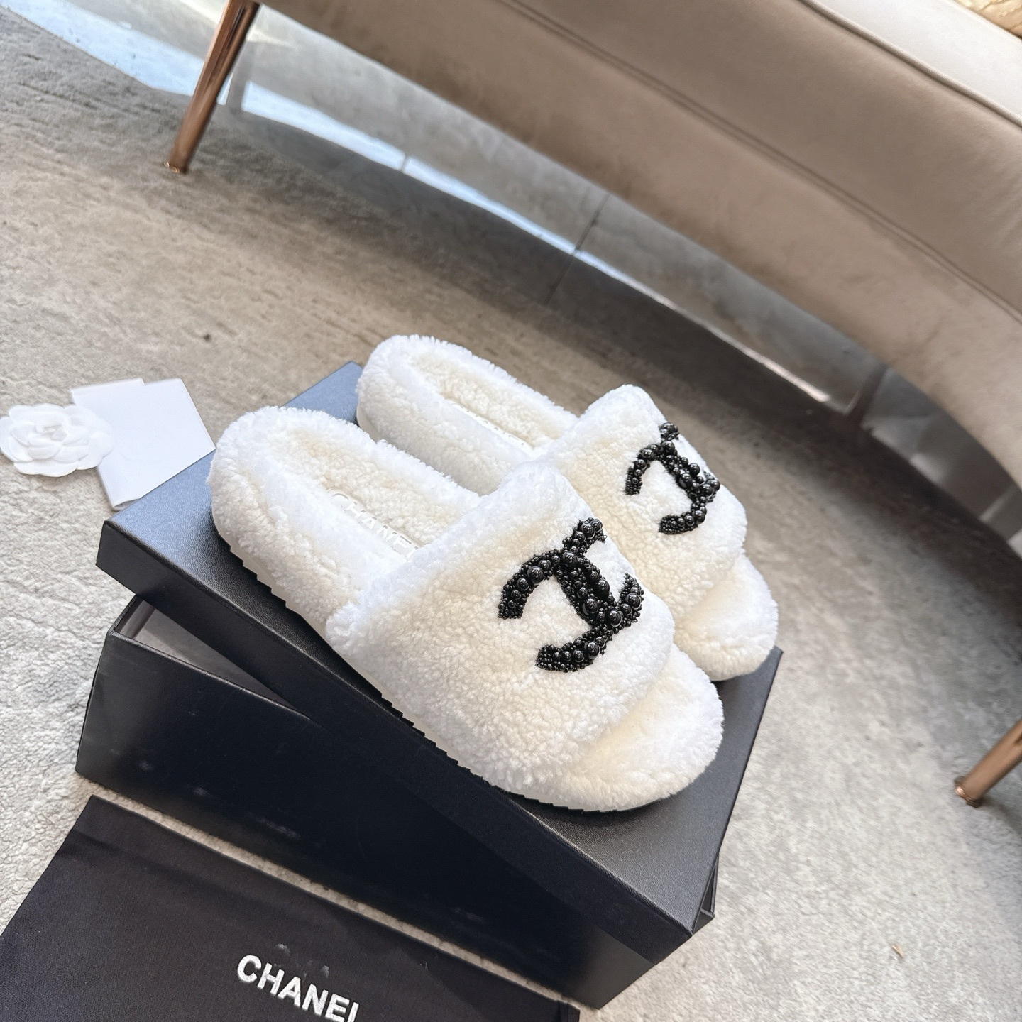 A pair of luxury Chanel white shearling slides featuring a black beaded CC logo, presented in a black Chanel box with a dust bag. - Hình 6