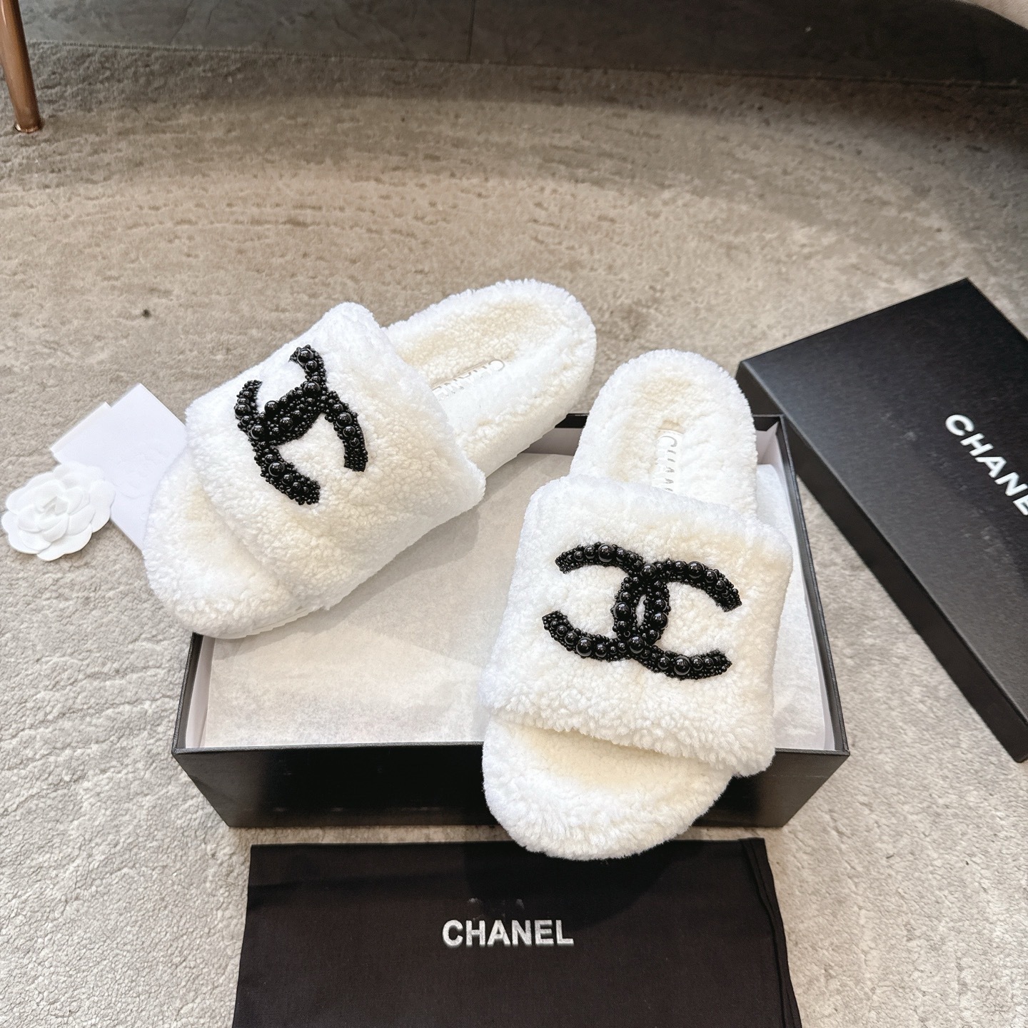 A pair of luxury Chanel white shearling slides featuring a black beaded CC logo, presented in a black Chanel box with a dust bag. - Hình 5