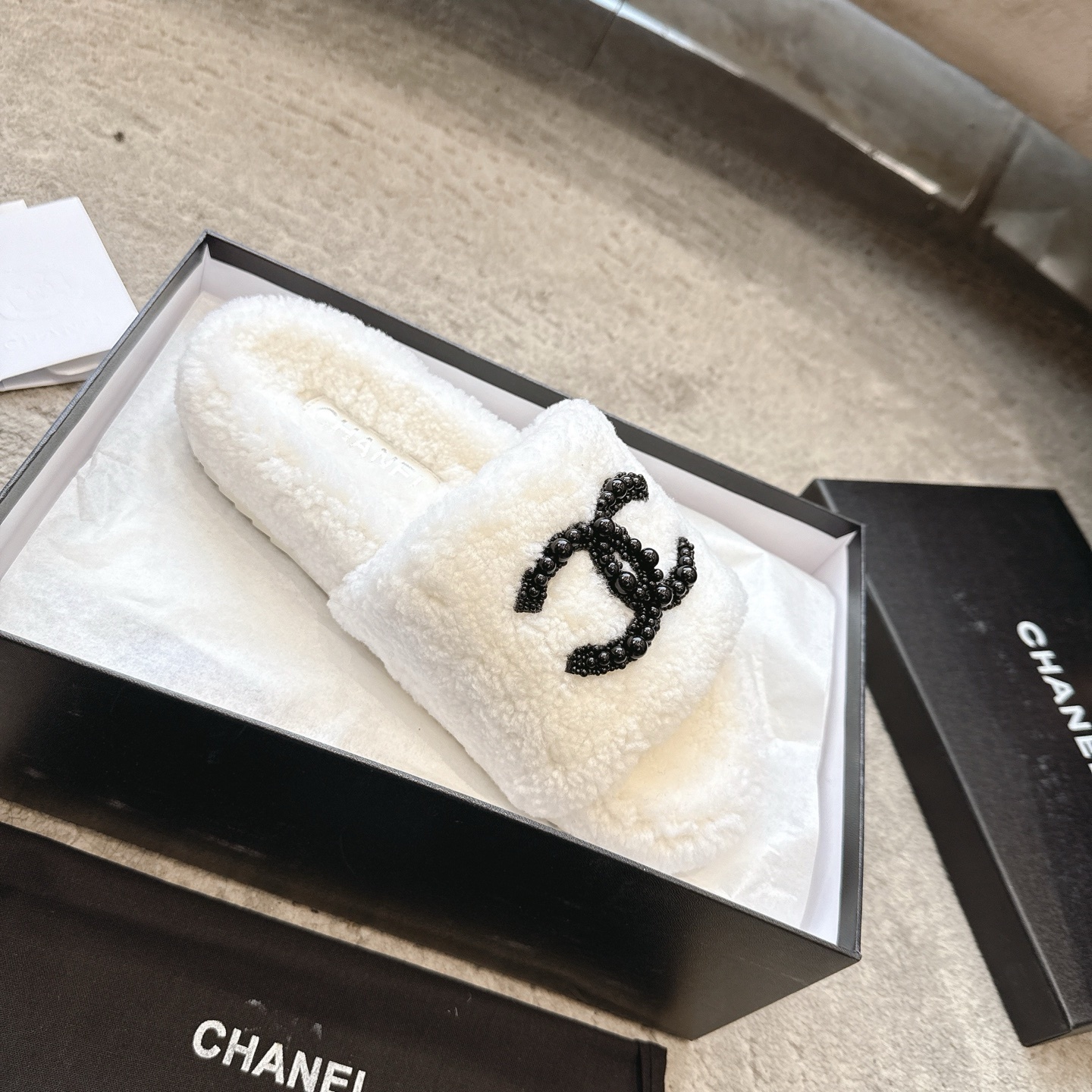 A pair of luxury Chanel white shearling slides featuring a black beaded CC logo, presented in a black Chanel box with a dust bag. - Hình 4