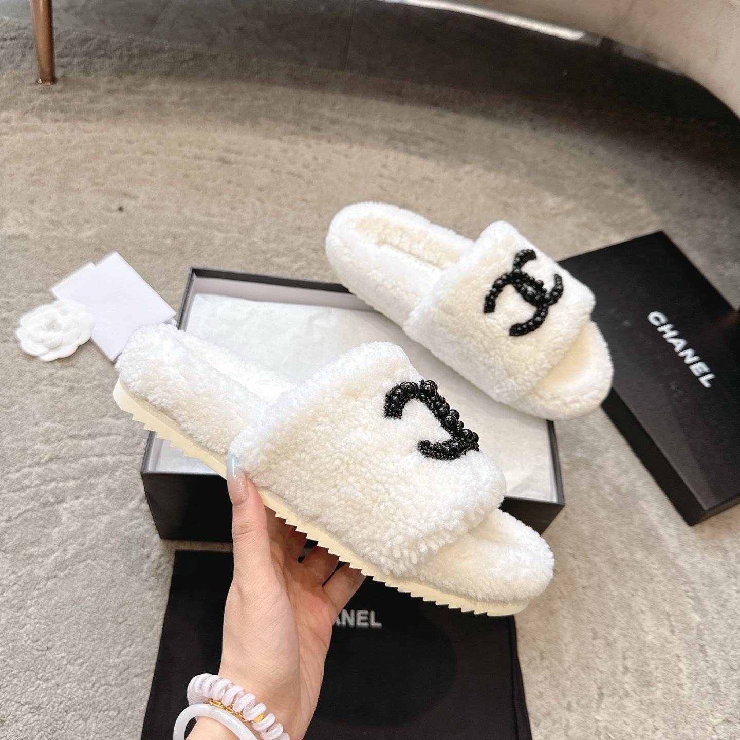 A pair of luxury Chanel white shearling slides featuring a black beaded CC logo, presented in a black Chanel box with a dust bag. - Hình 3