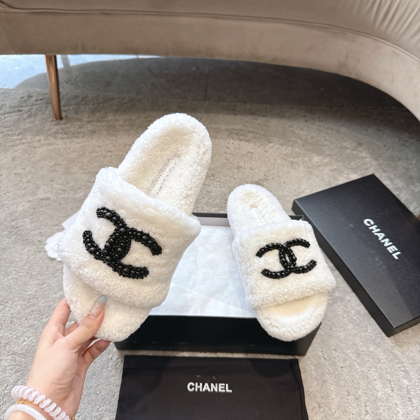 A pair of luxury Chanel white shearling slides featuring a black beaded CC logo, presented in a black Chanel box with a dust bag. - Hình 2