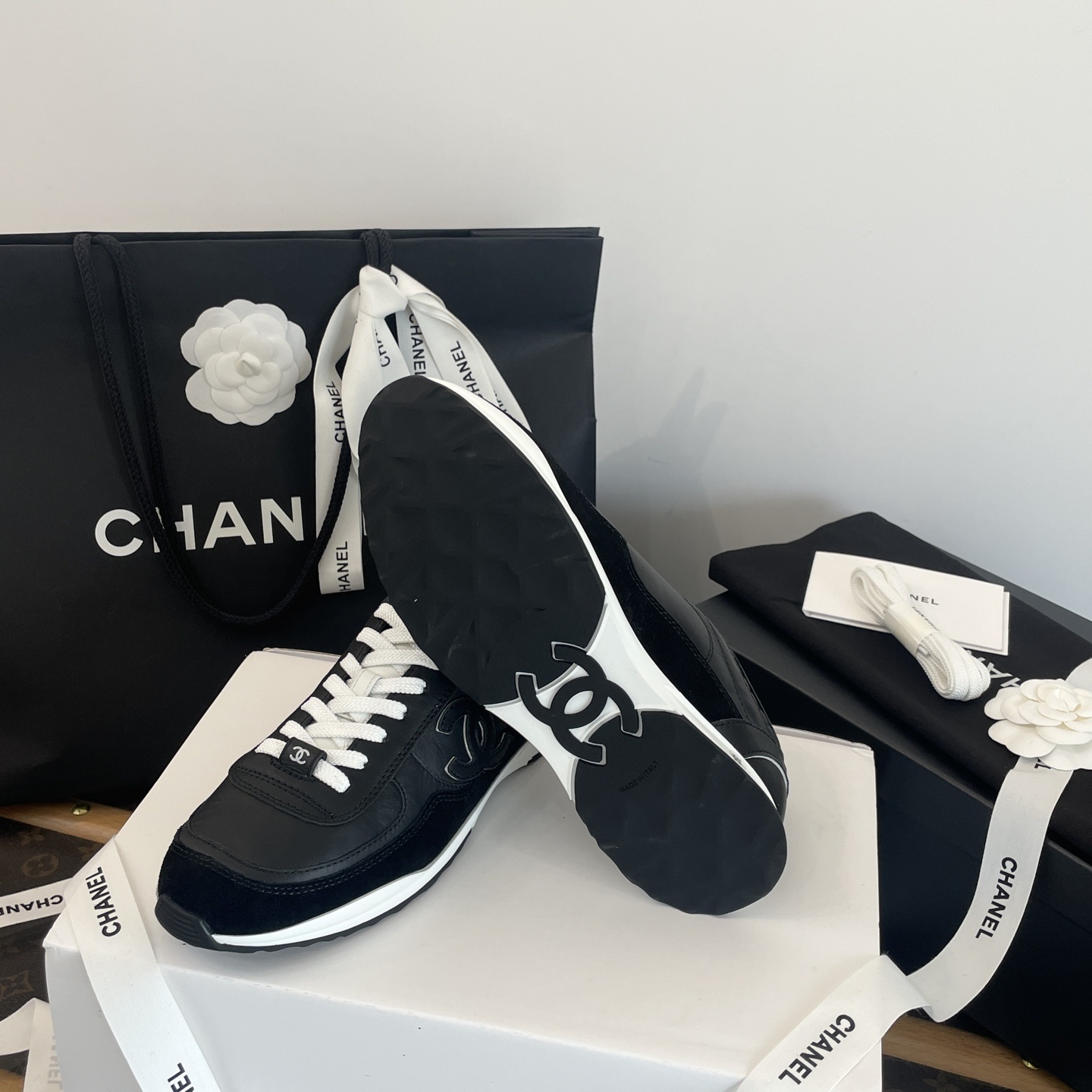 A pair of black Chanel leather sneakers with white laces displayed in original luxury packaging including a box and shopping bag. - Hình 9