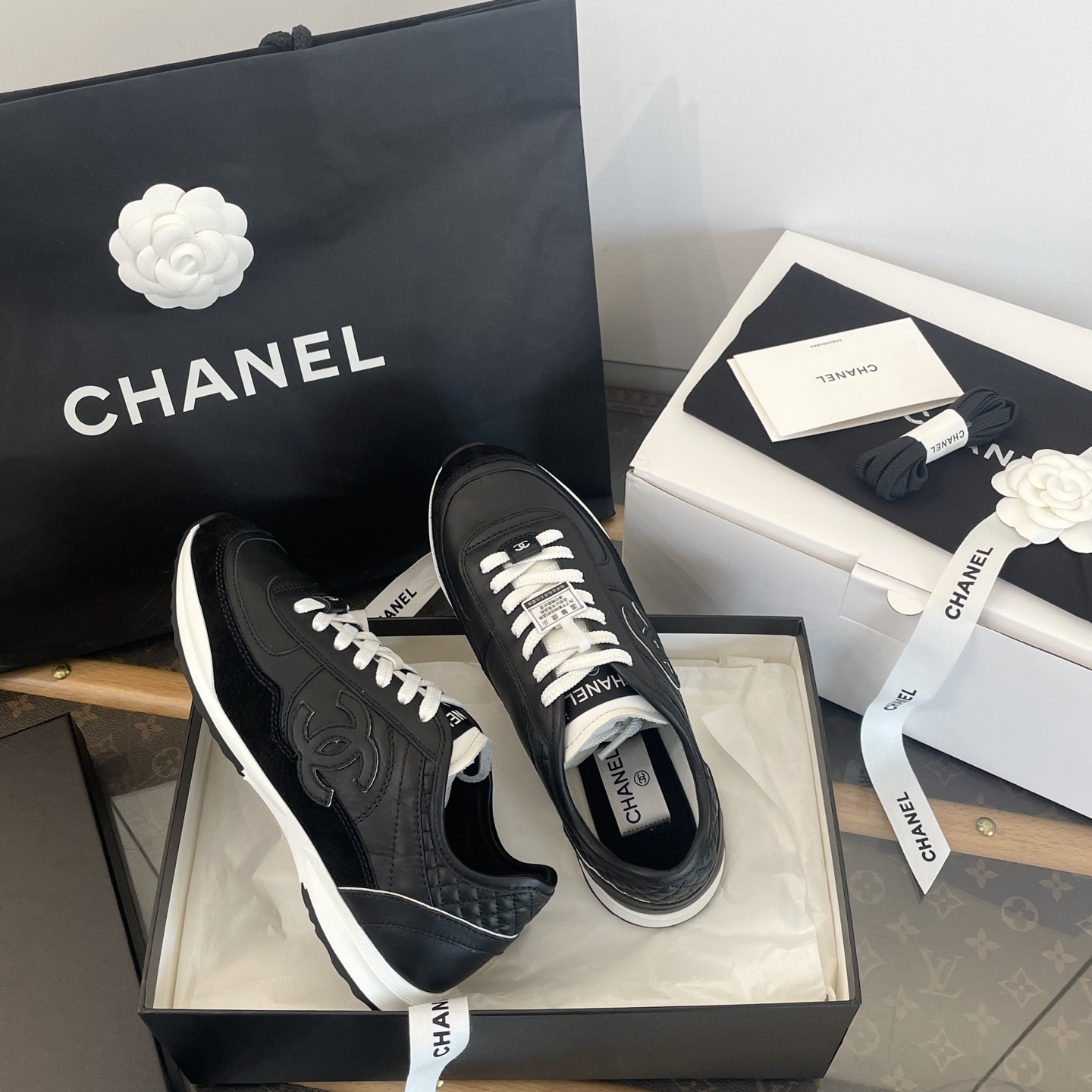 A pair of black Chanel leather sneakers with white laces displayed in original luxury packaging including a box and shopping bag. - Hình 8
