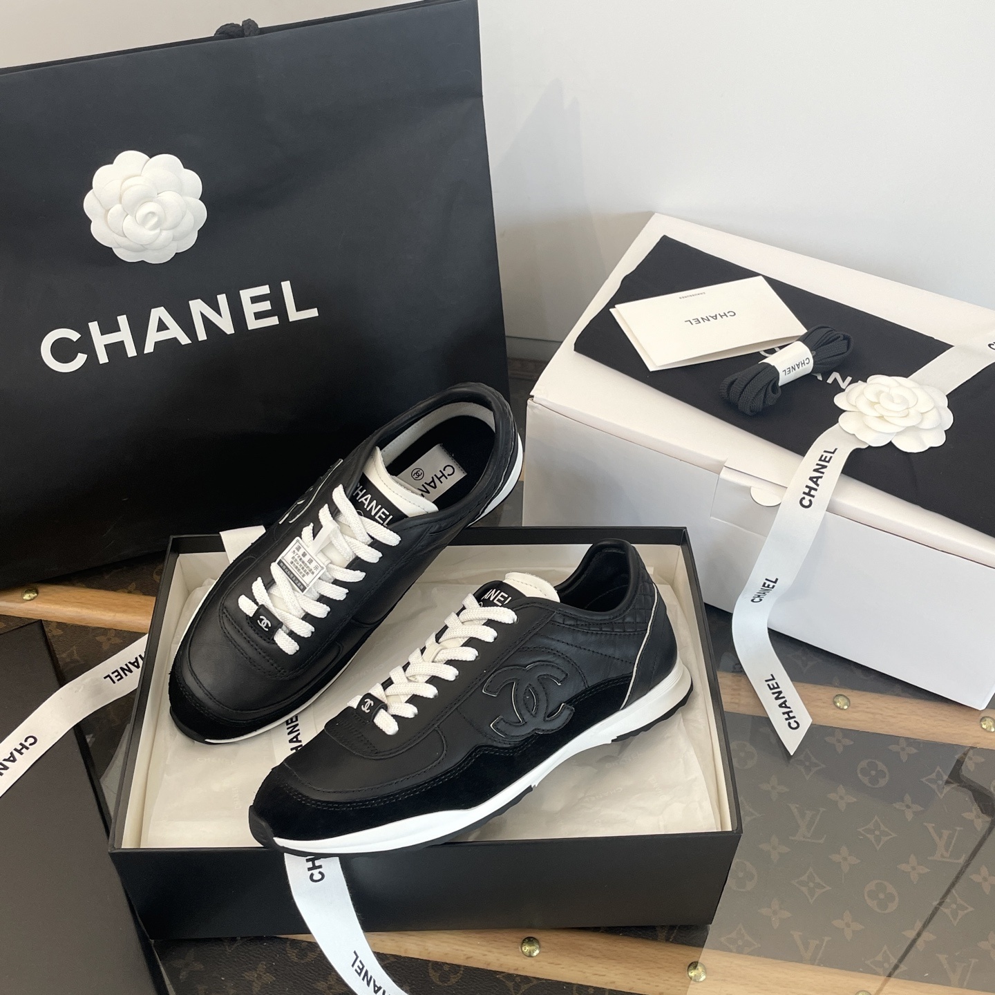 A pair of black Chanel leather sneakers with white laces displayed in original luxury packaging including a box and shopping bag. - Hình 6