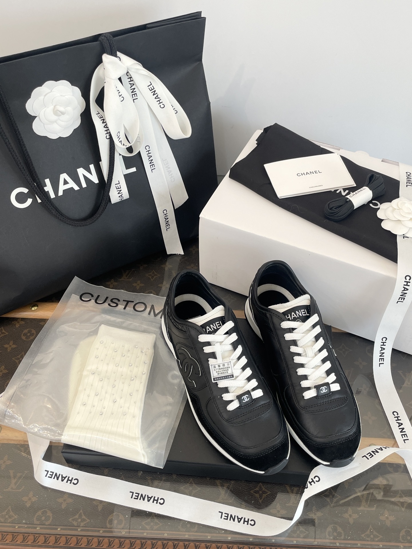 A pair of black Chanel leather sneakers with white laces displayed in original luxury packaging including a box and shopping bag. - Hình 5