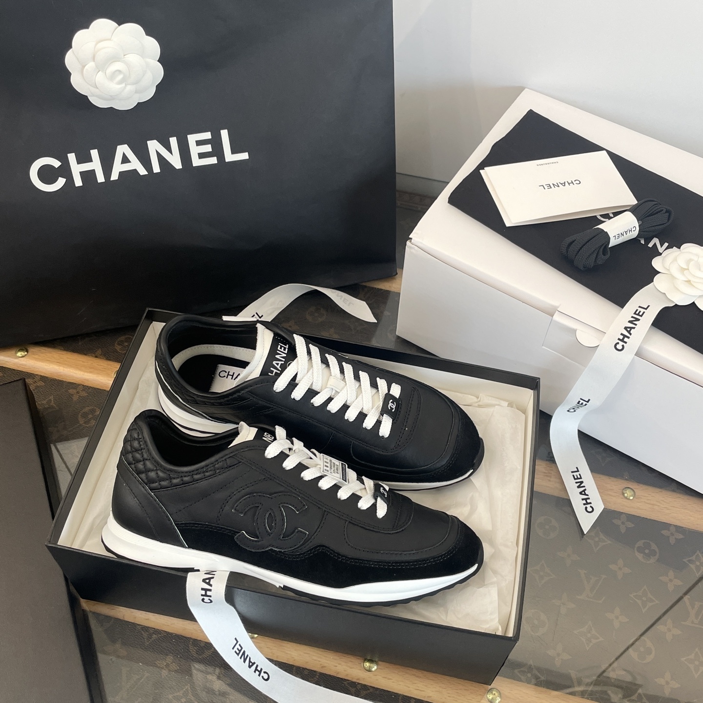 A pair of black Chanel leather sneakers with white laces displayed in original luxury packaging including a box and shopping bag. - Hình 4