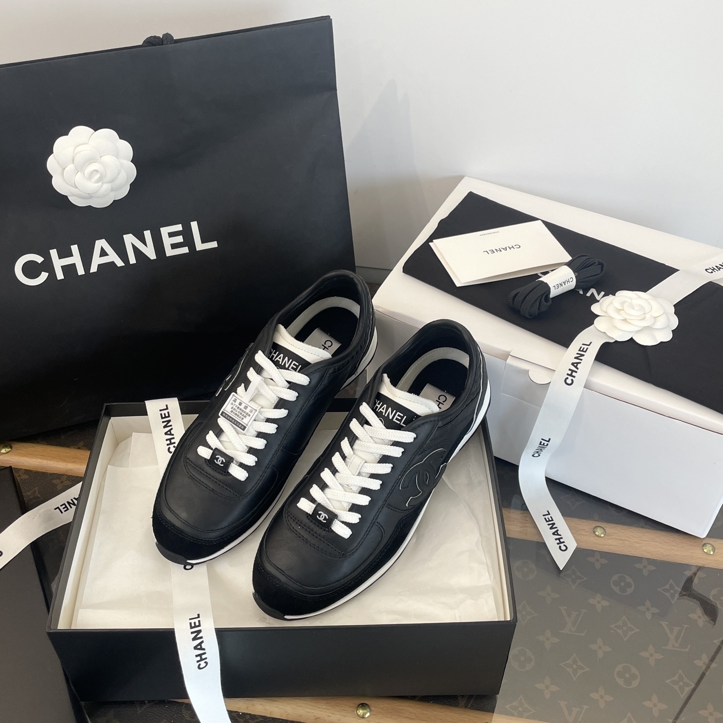A pair of black Chanel leather sneakers with white laces displayed in original luxury packaging including a box and shopping bag. - Hình 3