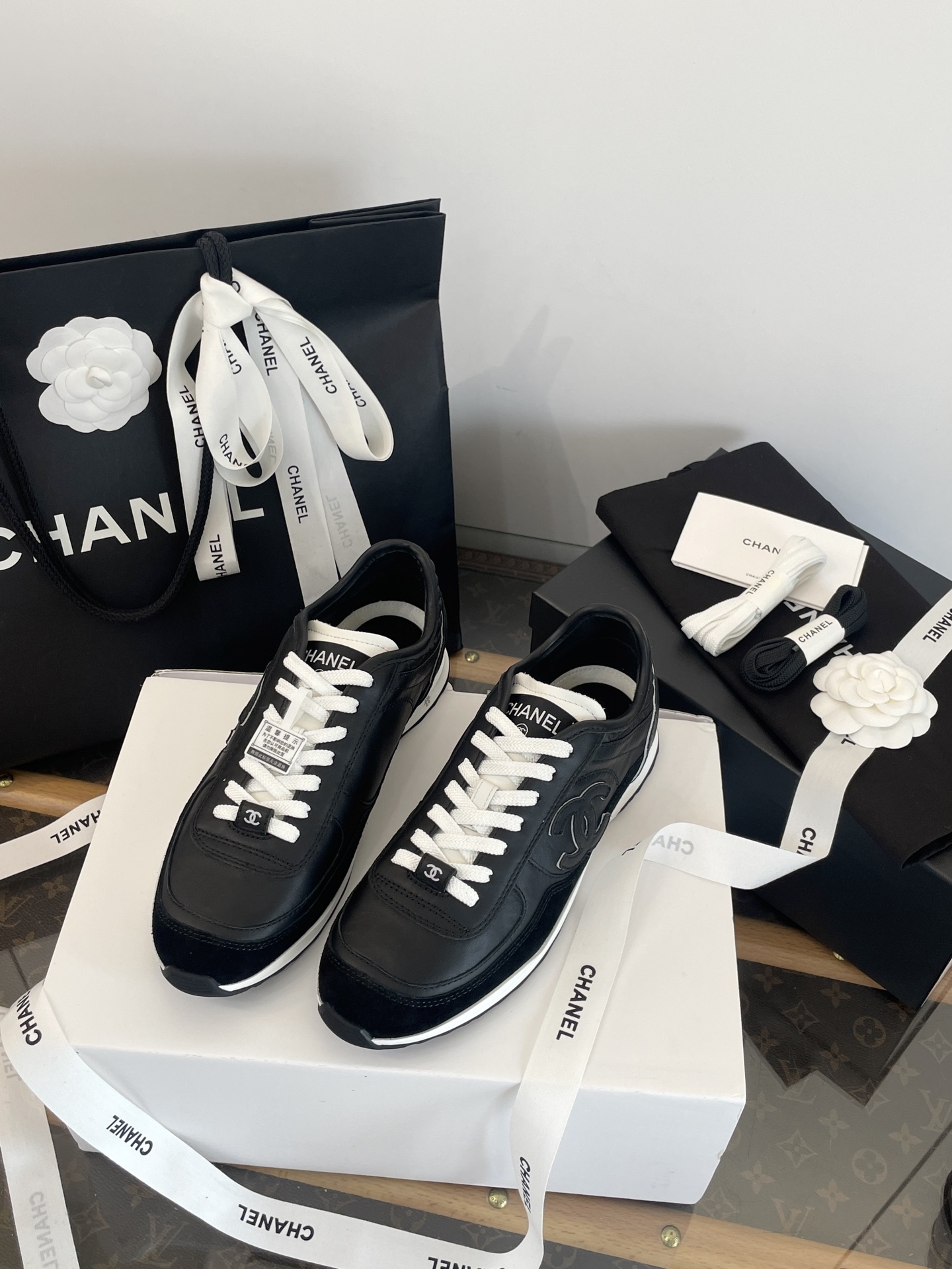 A pair of black Chanel leather sneakers with white laces displayed in original luxury packaging including a box and shopping bag. - Hình 2