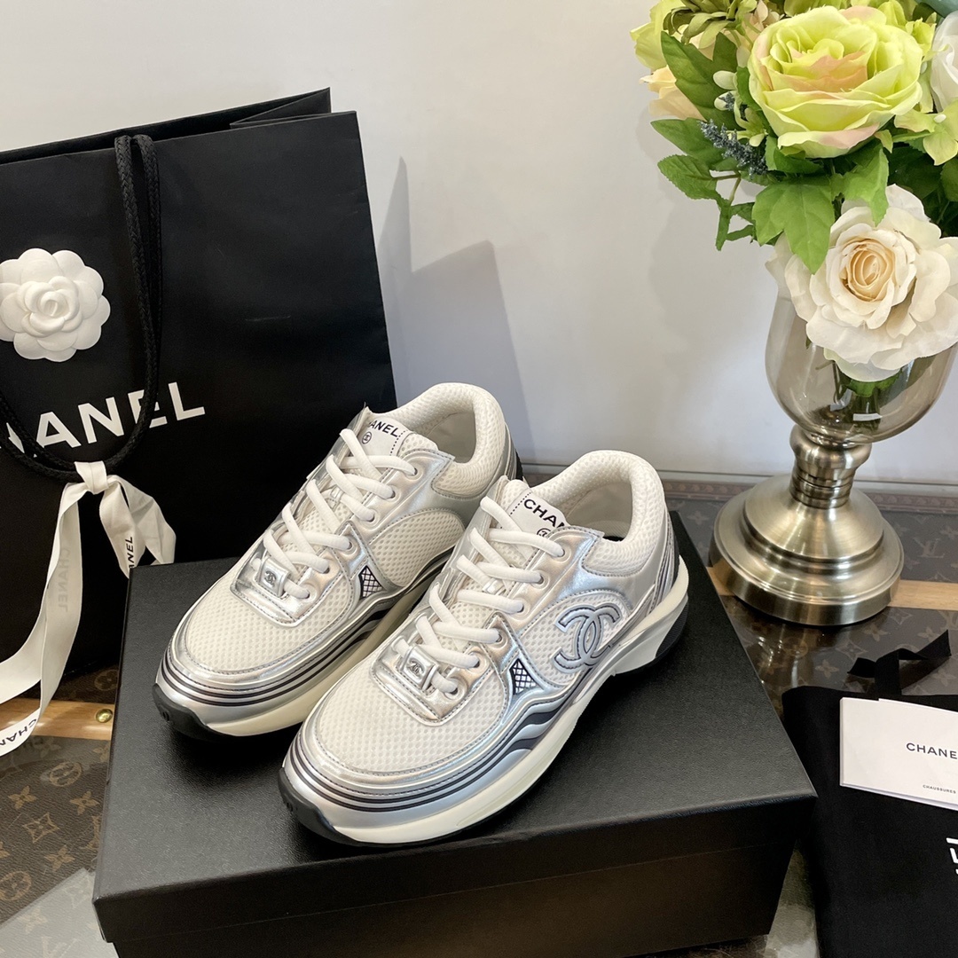 Pair of Chanel metallic silver and white mesh sneakers on a black box with a luxury shopping bag. - Hình 2