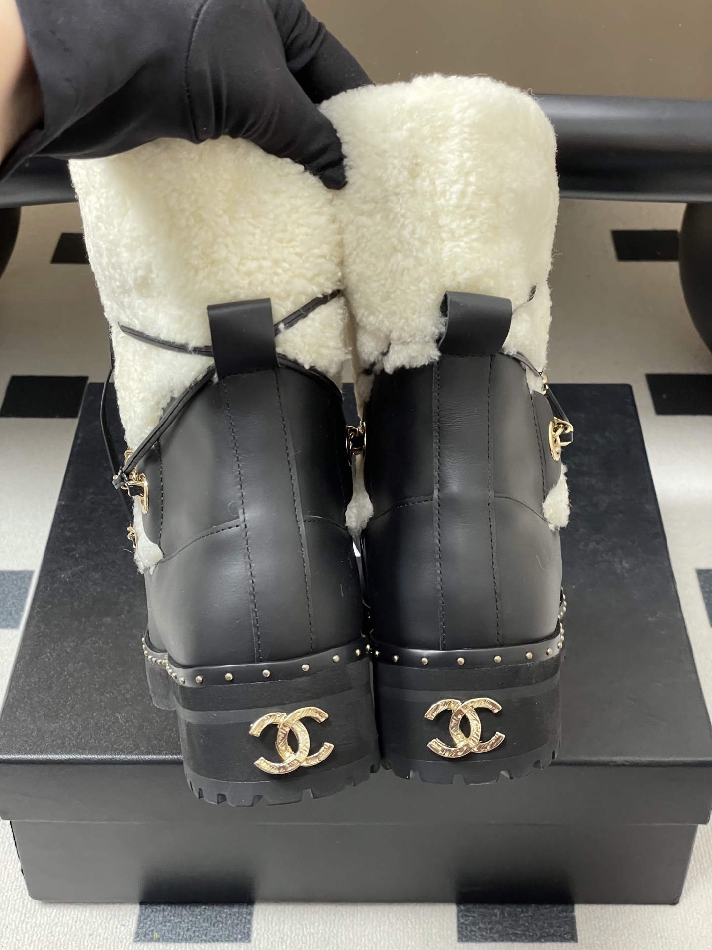 Pair of luxury black leather and white shearling combat boots with gold chain accents and embossed logo on a black table. - Hình 10