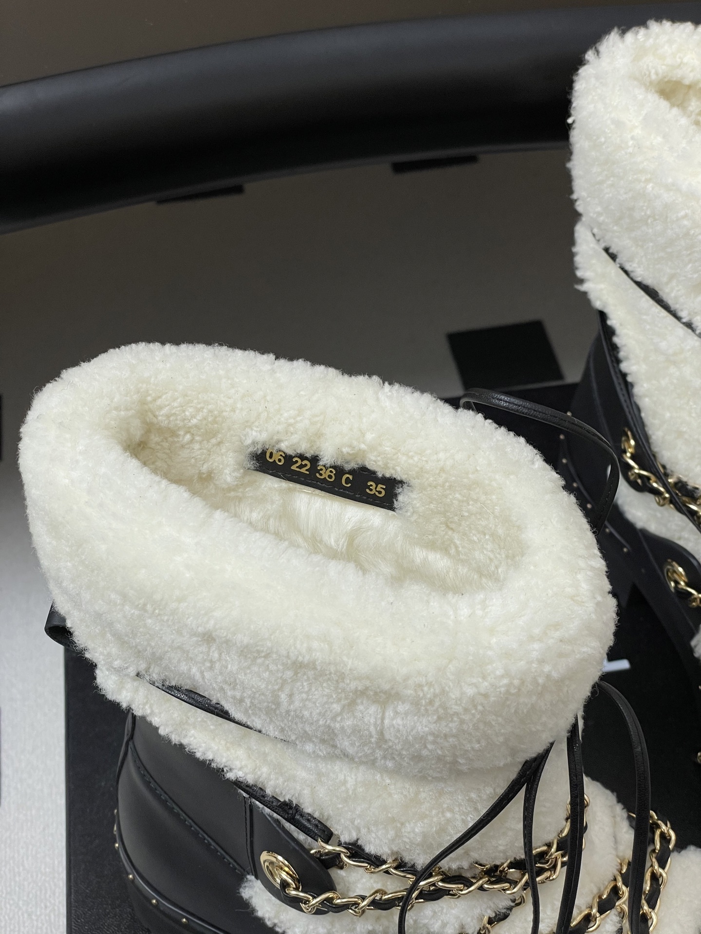 Pair of luxury black leather and white shearling combat boots with gold chain accents and embossed logo on a black table. - Hình 9