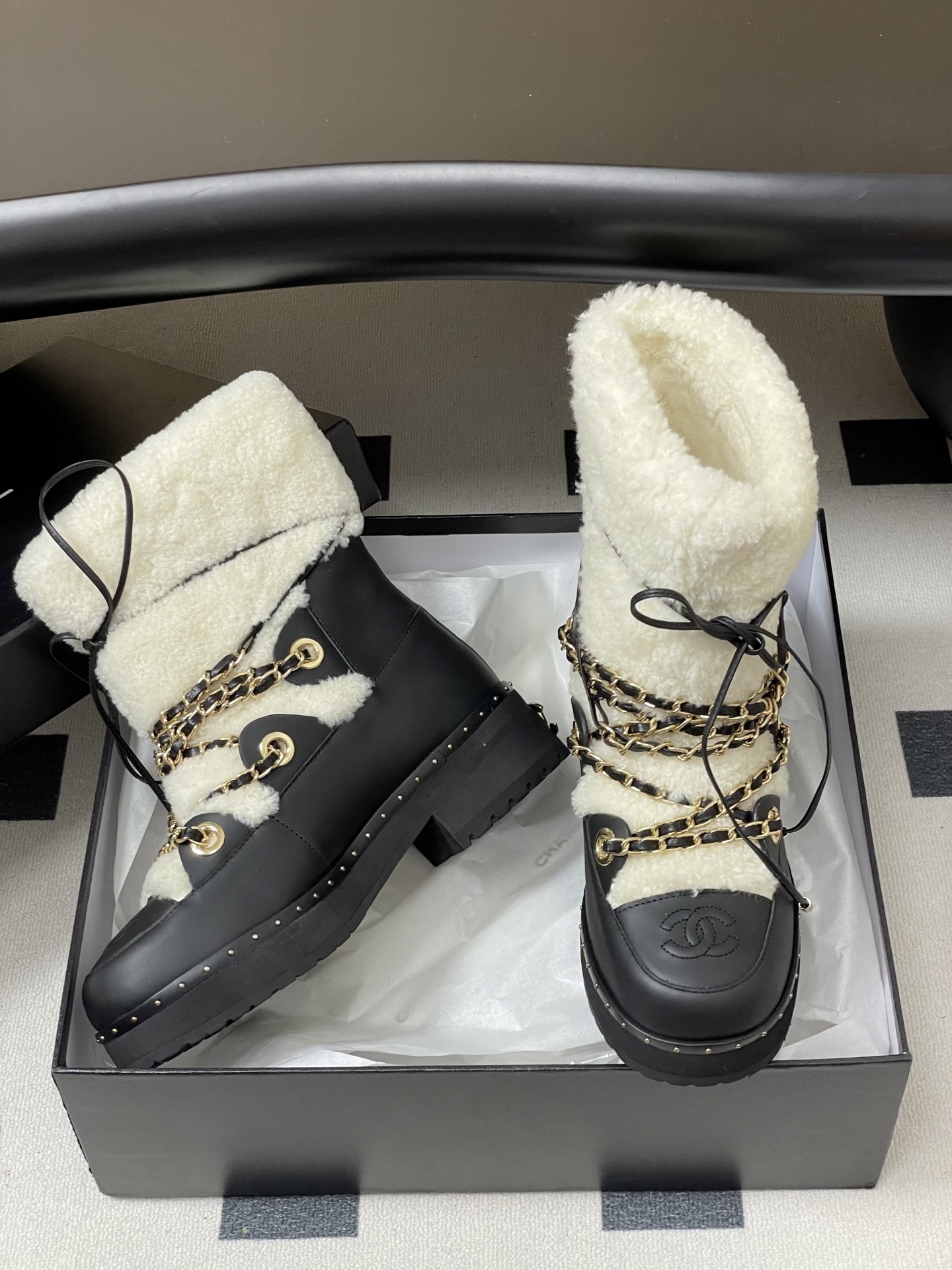 Pair of luxury black leather and white shearling combat boots with gold chain accents and embossed logo on a black table. - Hình 7