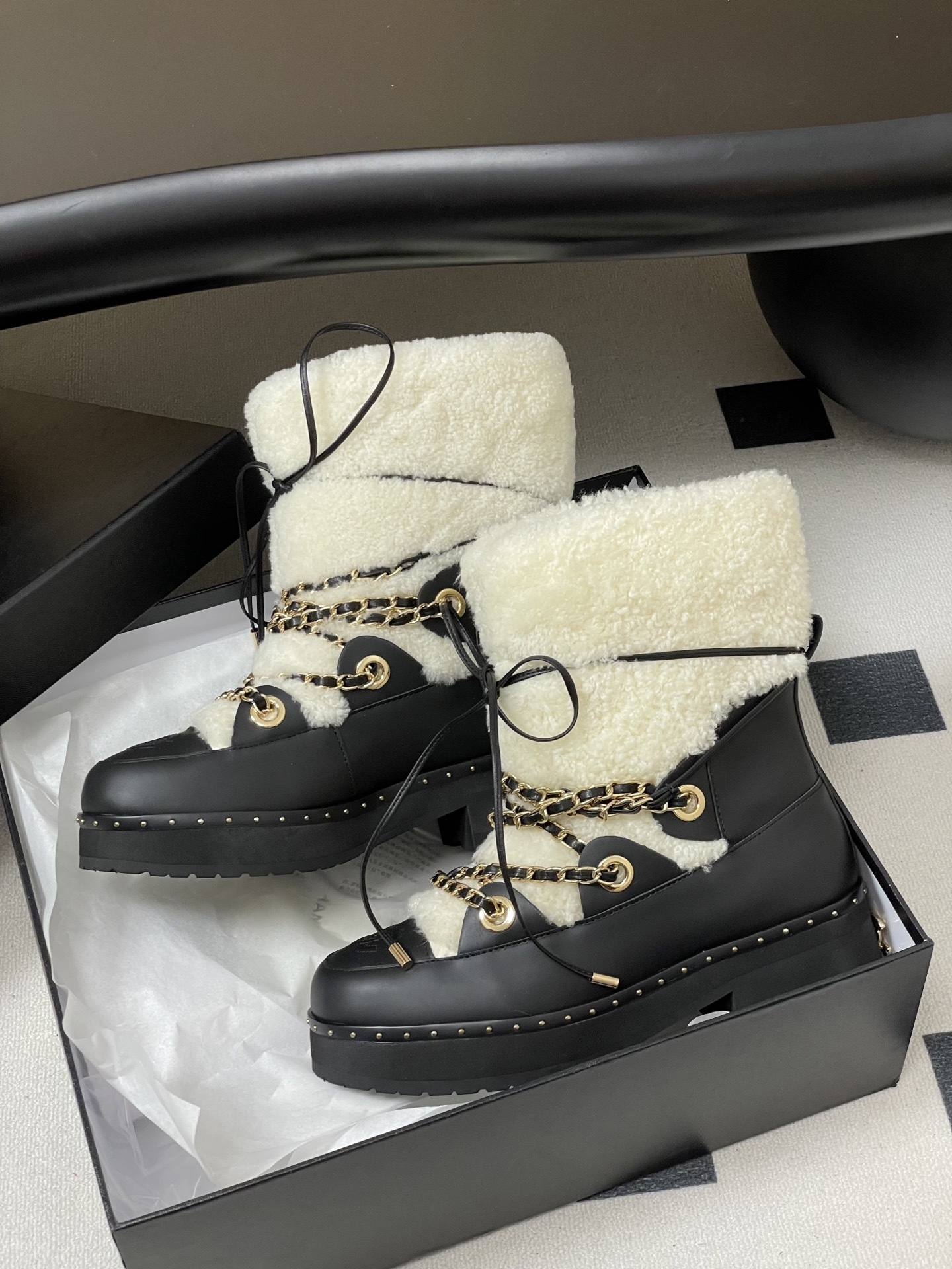 Pair of luxury black leather and white shearling combat boots with gold chain accents and embossed logo on a black table. - Hình 5