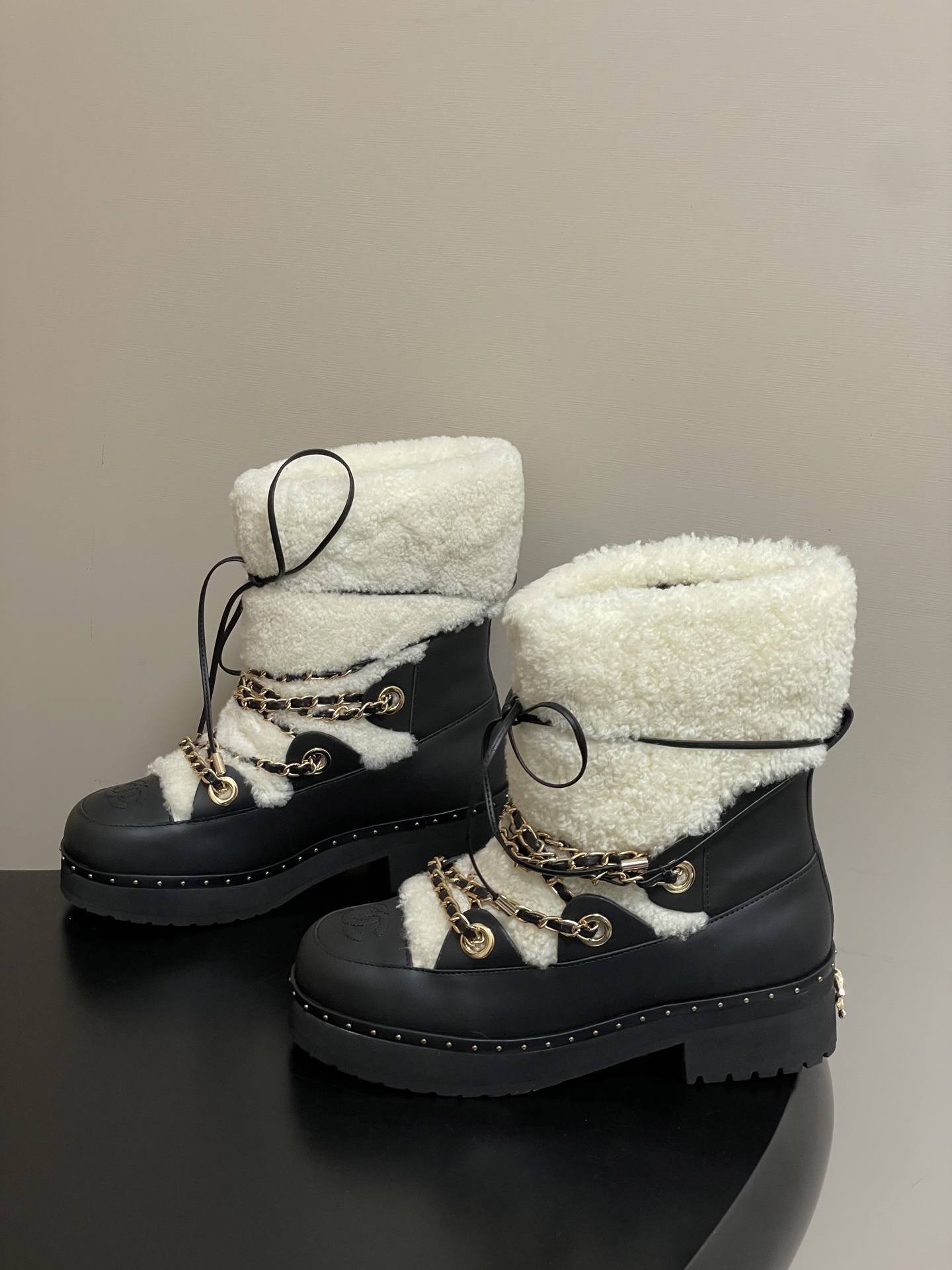 Pair of luxury black leather and white shearling combat boots with gold chain accents and embossed logo on a black table. - Hình 4