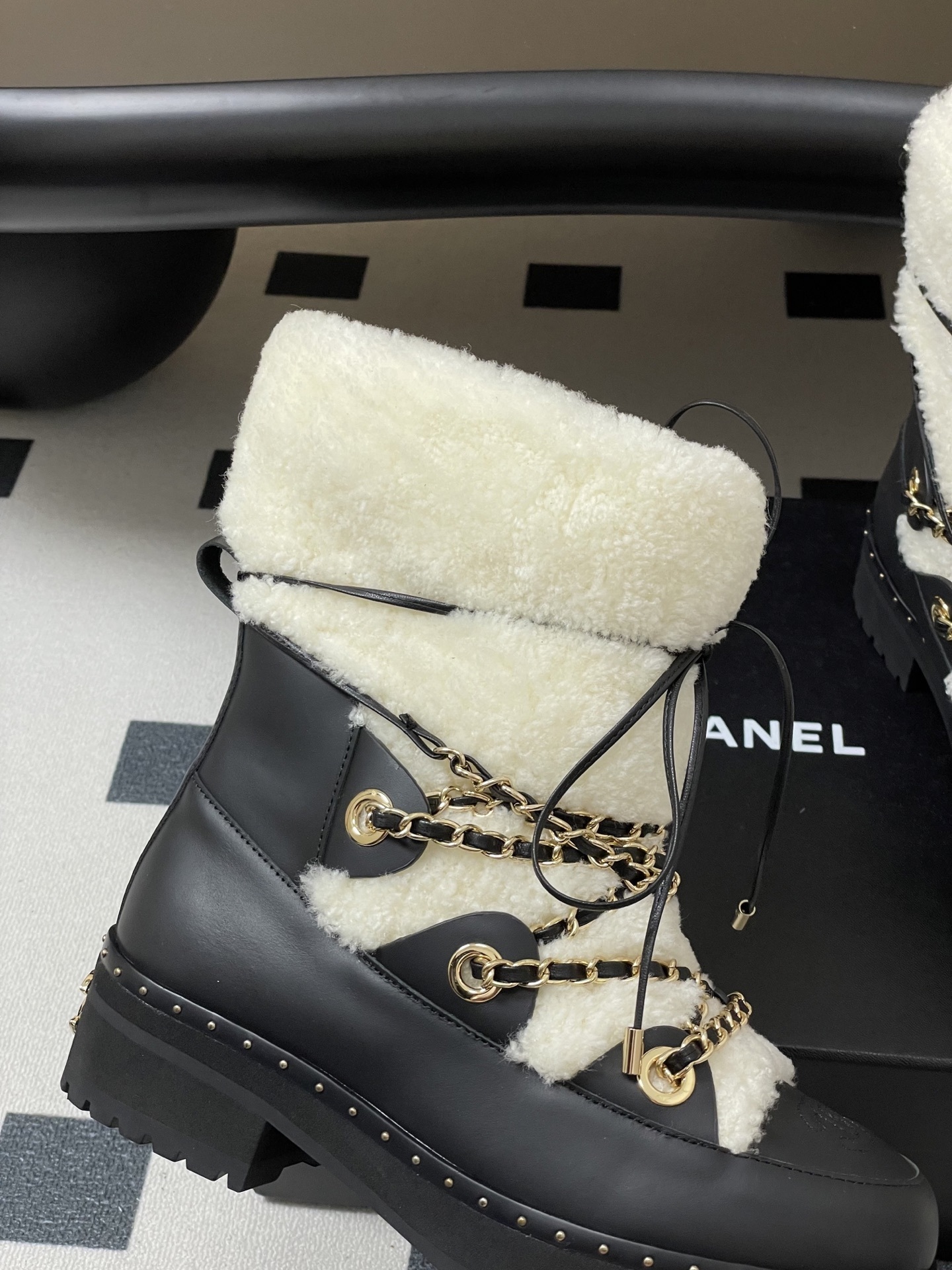 Pair of luxury black leather and white shearling combat boots with gold chain accents and embossed logo on a black table. - Hình 2