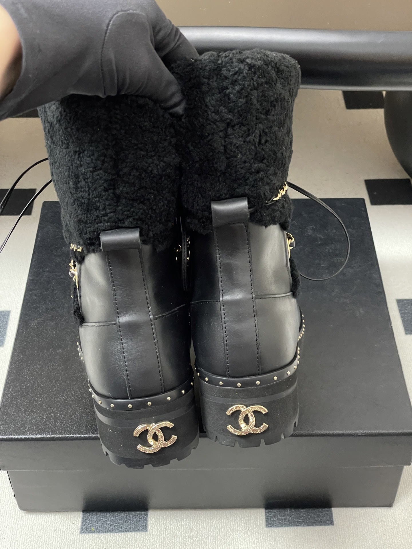 Pair of black leather combat boots with black shearling trim, gold chain detailing, and lace-up front on a black pedestal. - Hình 8