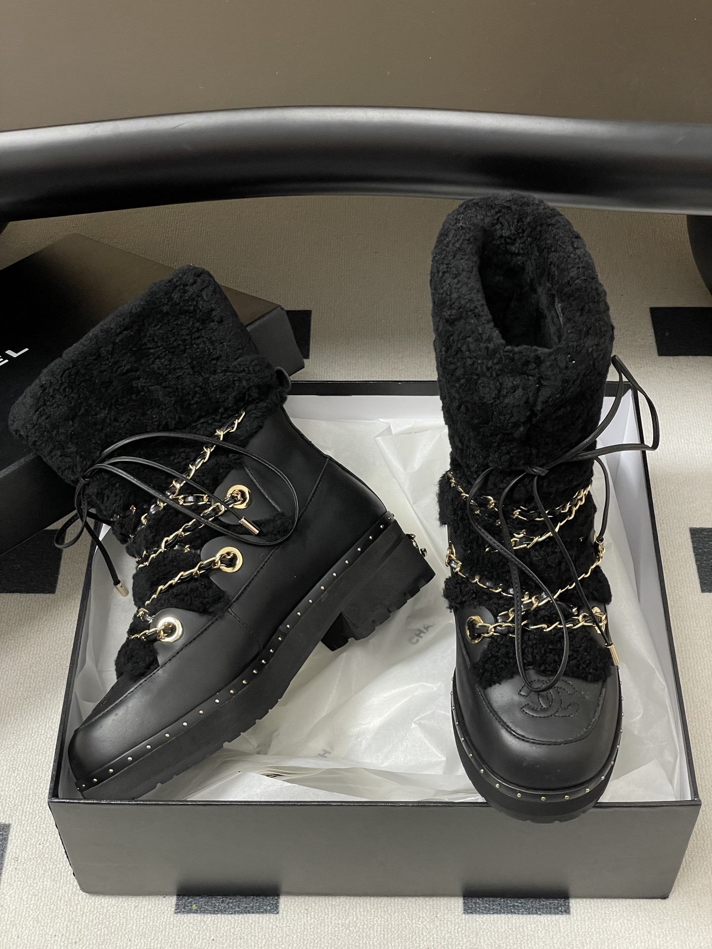 Pair of black leather combat boots with black shearling trim, gold chain detailing, and lace-up front on a black pedestal. - Hình 7