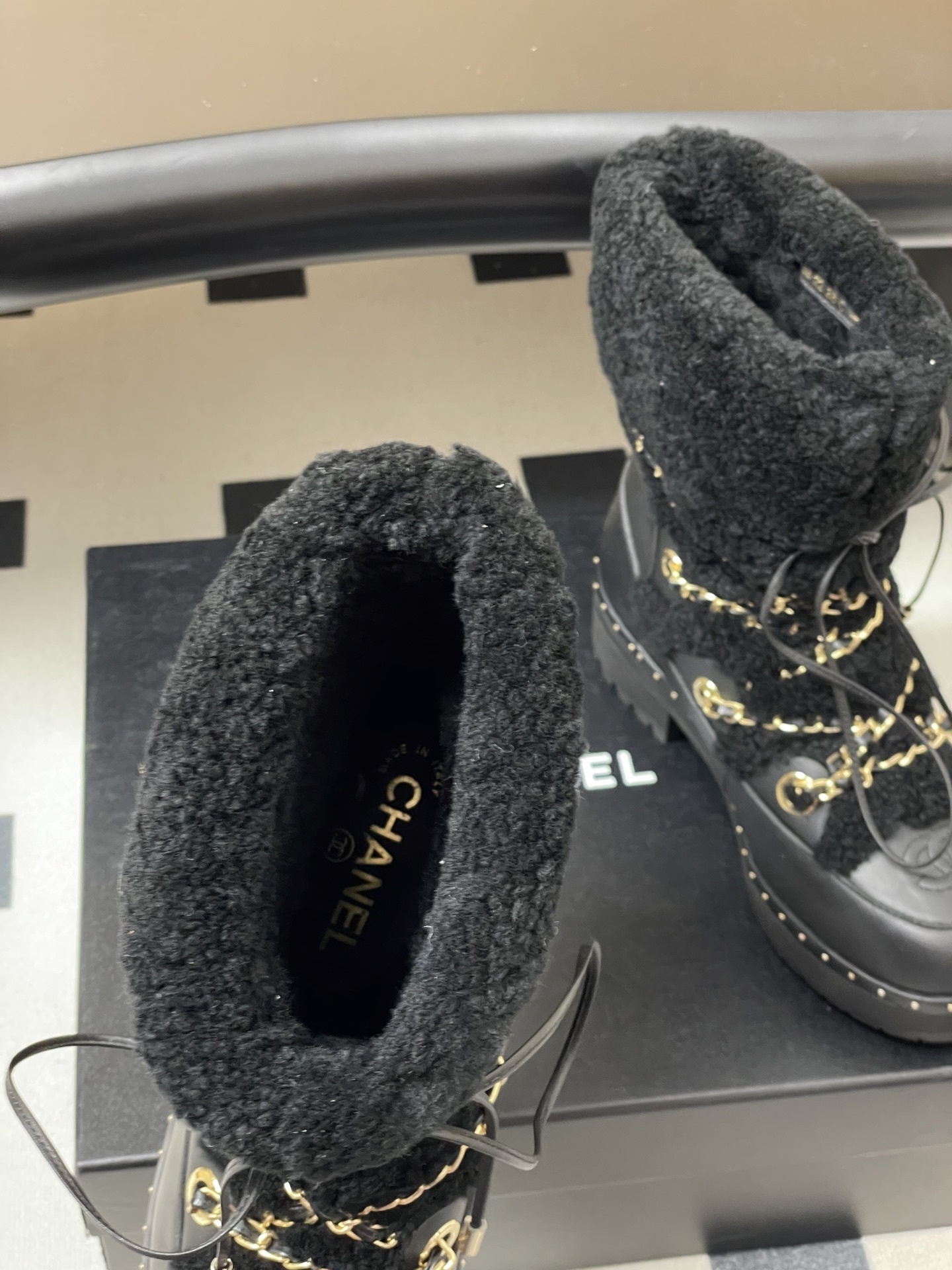 Pair of black leather combat boots with black shearling trim, gold chain detailing, and lace-up front on a black pedestal. - Hình 6