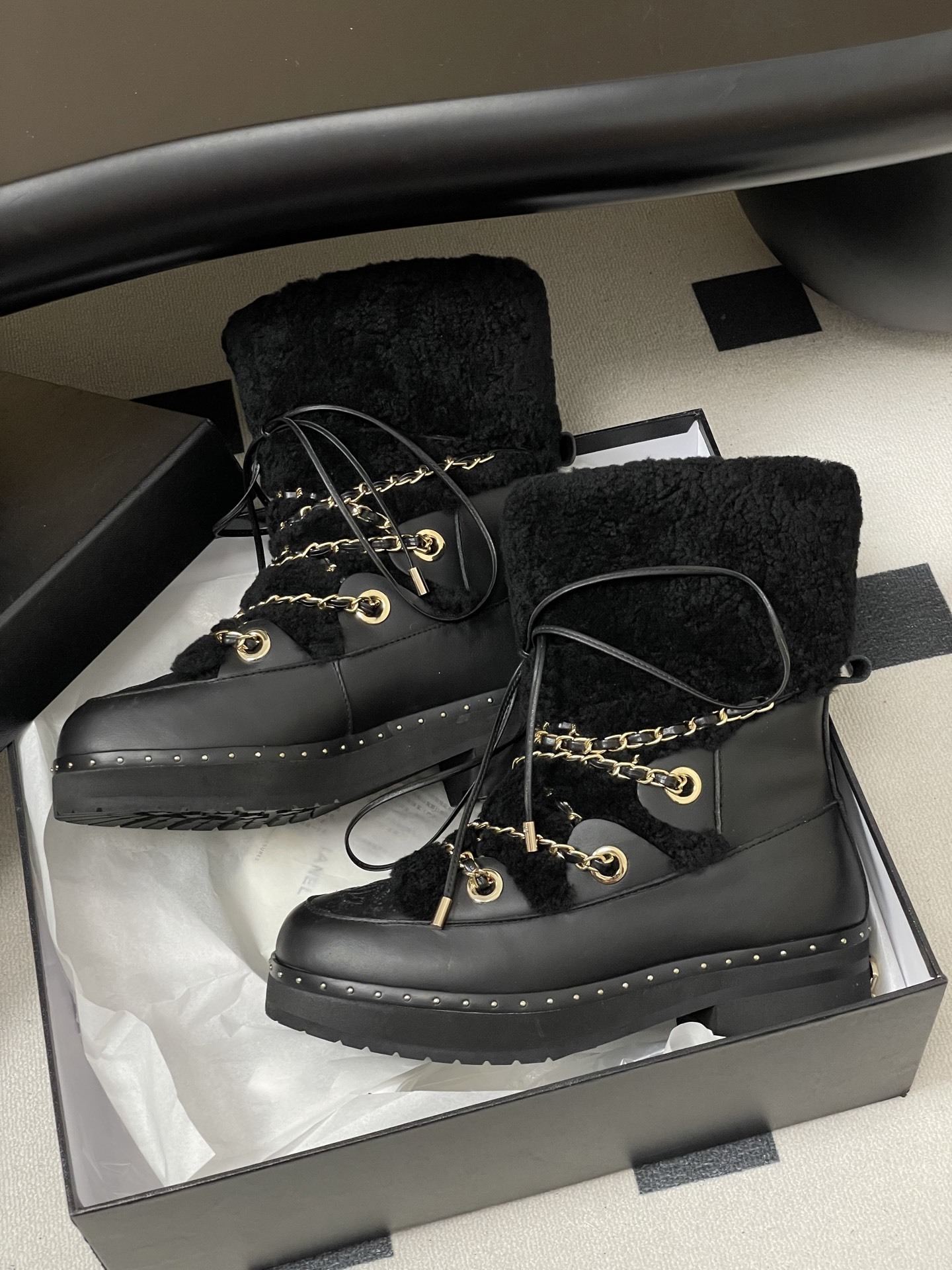Pair of black leather combat boots with black shearling trim, gold chain detailing, and lace-up front on a black pedestal. - Hình 5
