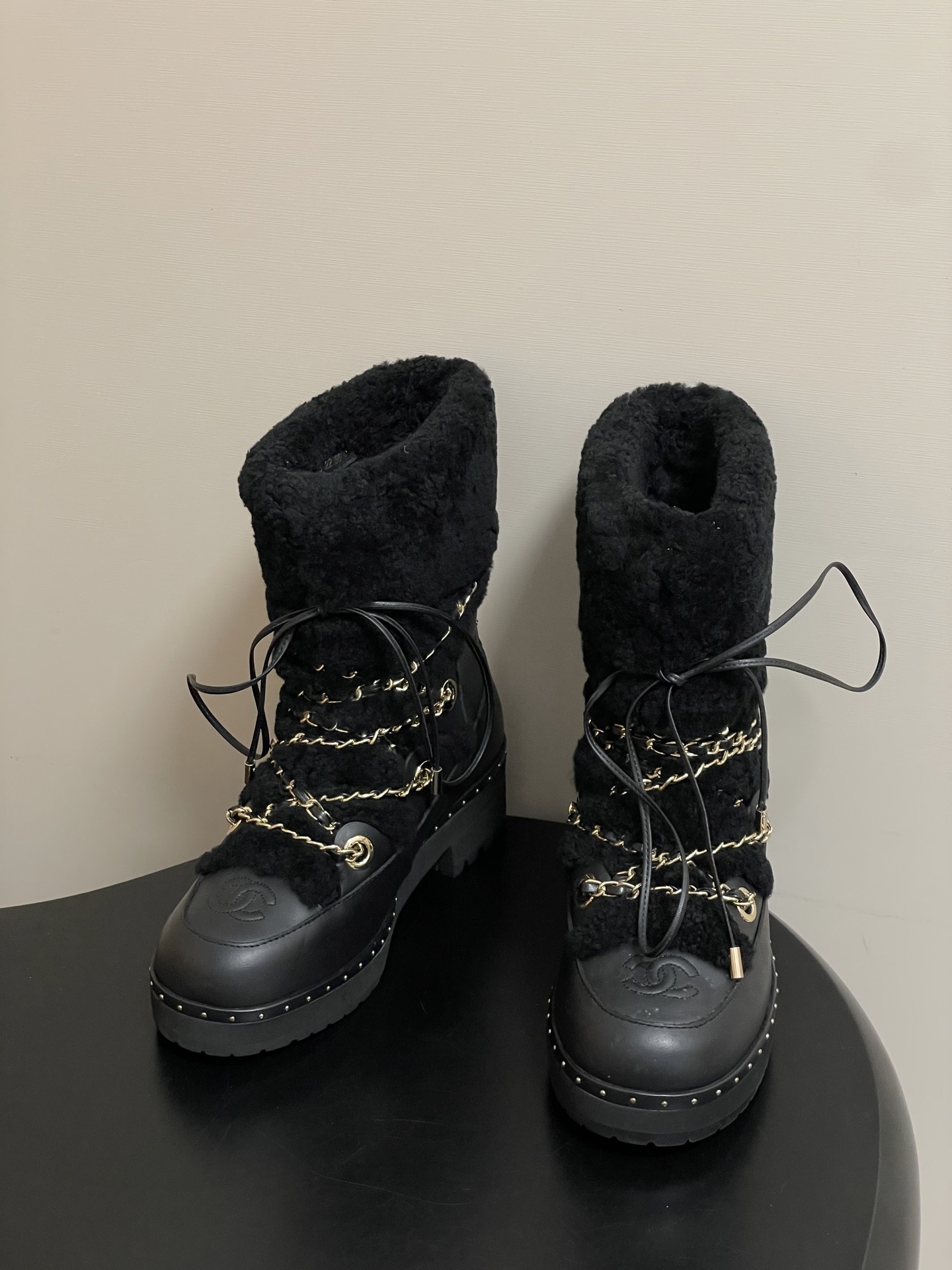 Pair of black leather combat boots with black shearling trim, gold chain detailing, and lace-up front on a black pedestal. - Hình 3